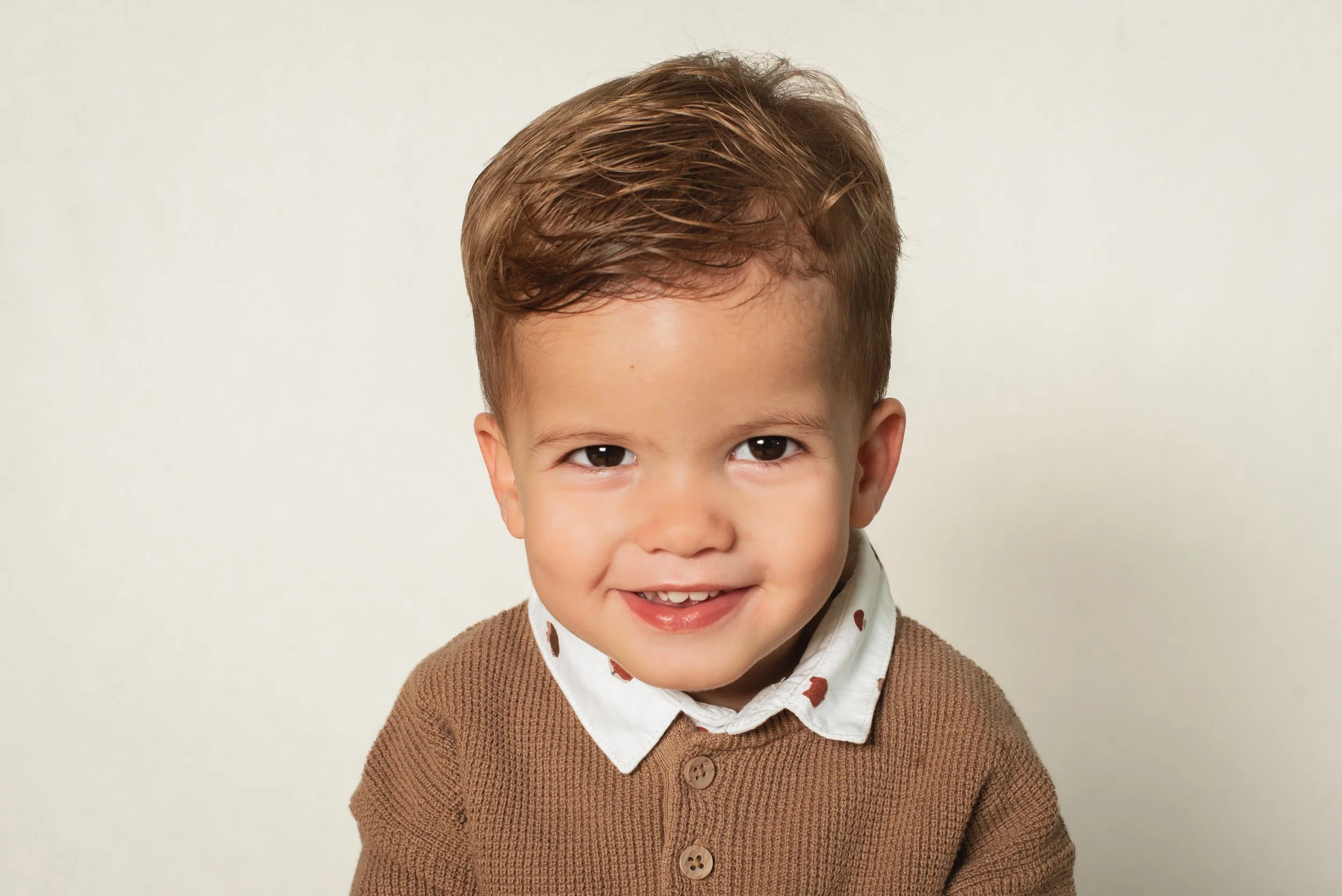 A young boy with brown hair wearing a brown sweater and a white shirt with a pattern, smiling at the camera against a plain light-colored background.