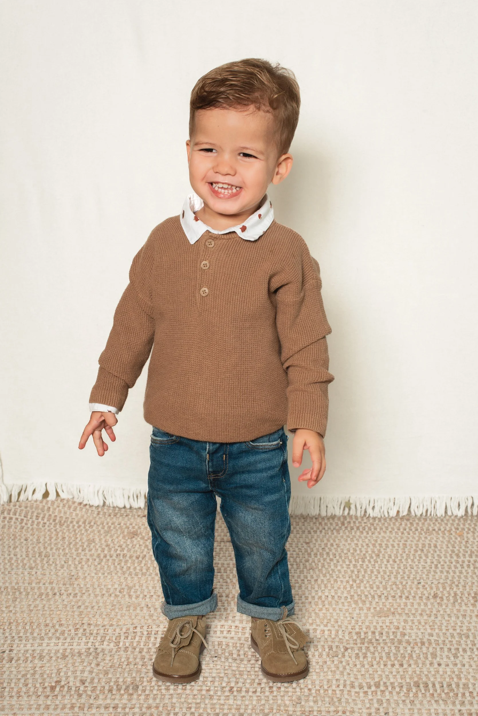 A young boy with a big smile, wearing a brown sweater, jeans, and beige boots, standing indoors on a beige textured rug, against a plain white wall.