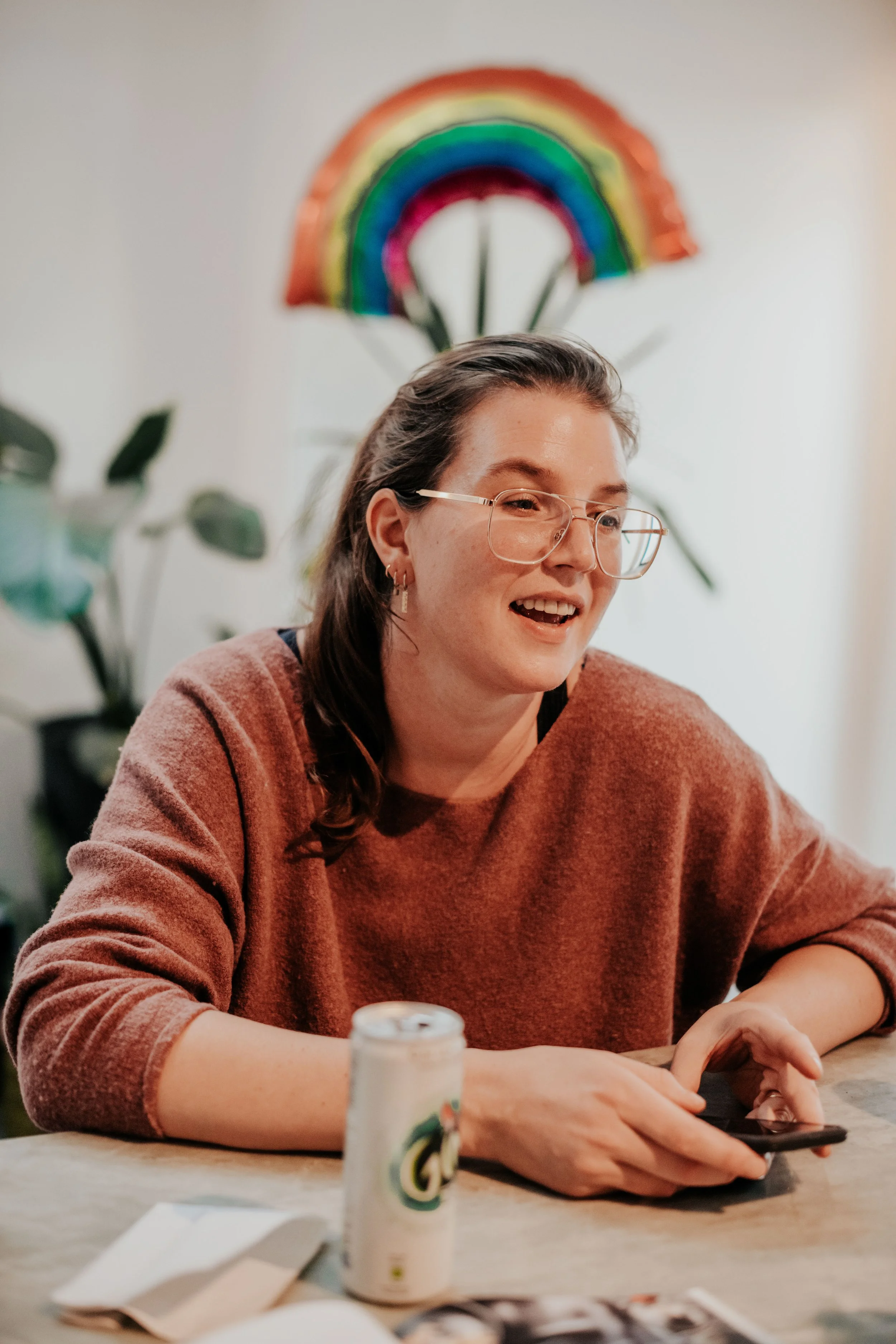 A woman with glasses and earrings, wearing a brown sweater, sitting at a table with a phone in her hands. A rainbow decoration is behind her on the wall, with green leaves of a plant visible on the side. A glue stick, paper, and photograph are on the