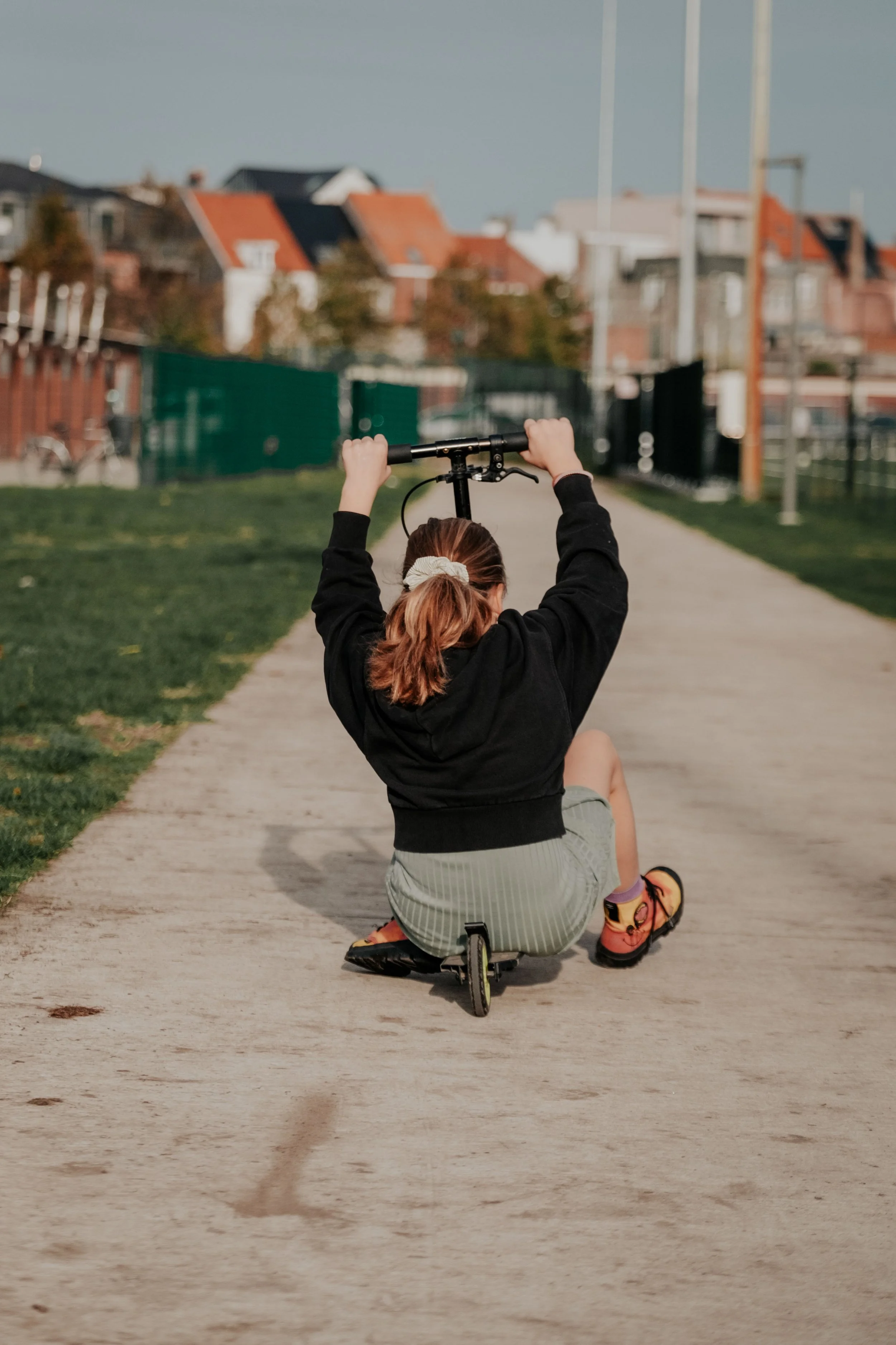 A person with red hair sitting on a small scooter, holding a handlebar above their head, on a pathway in a park.