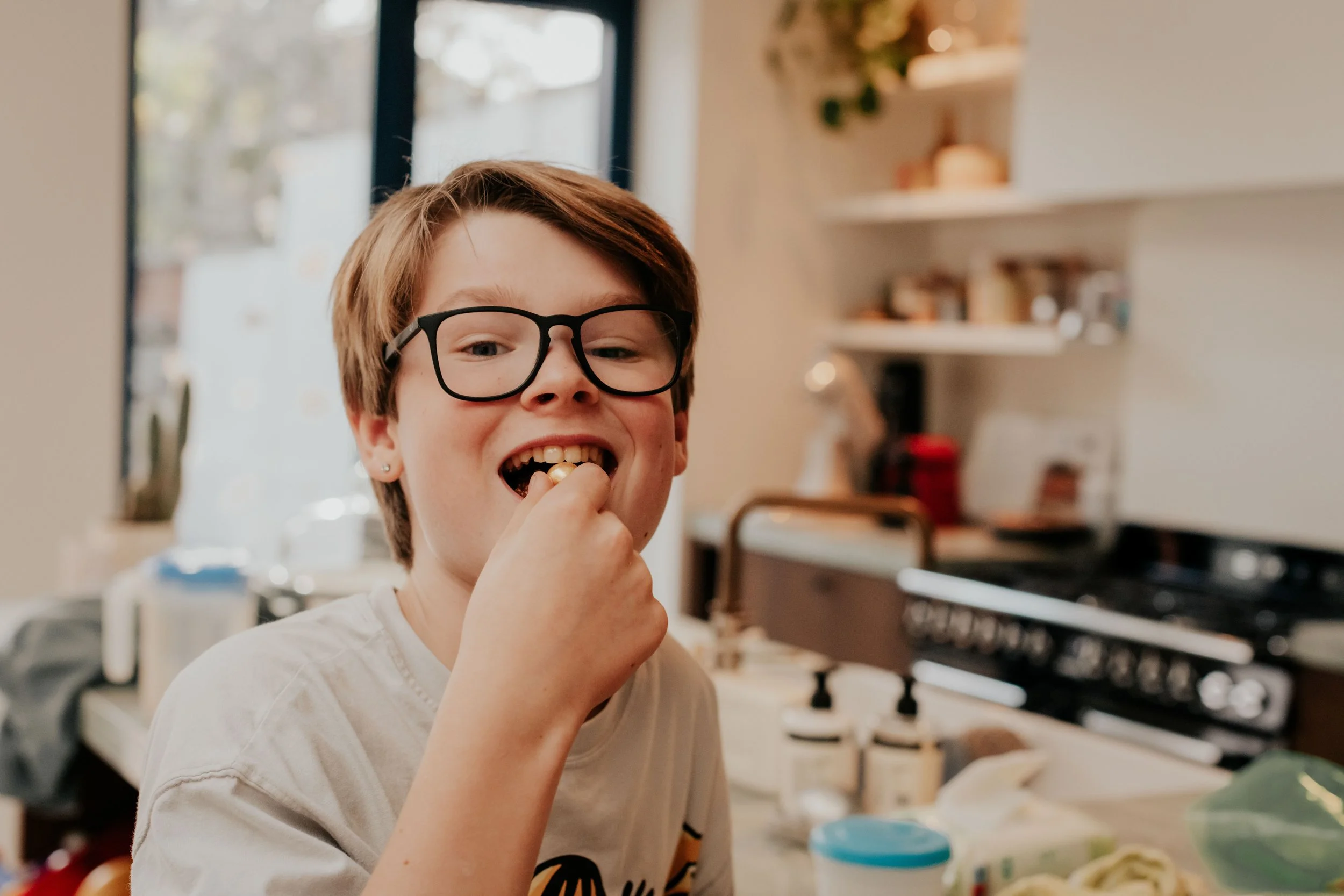 A young person with short brown hair, glasses, and earrings, smiling while eating a snack in a kitchen with shelves and a stove in the background.