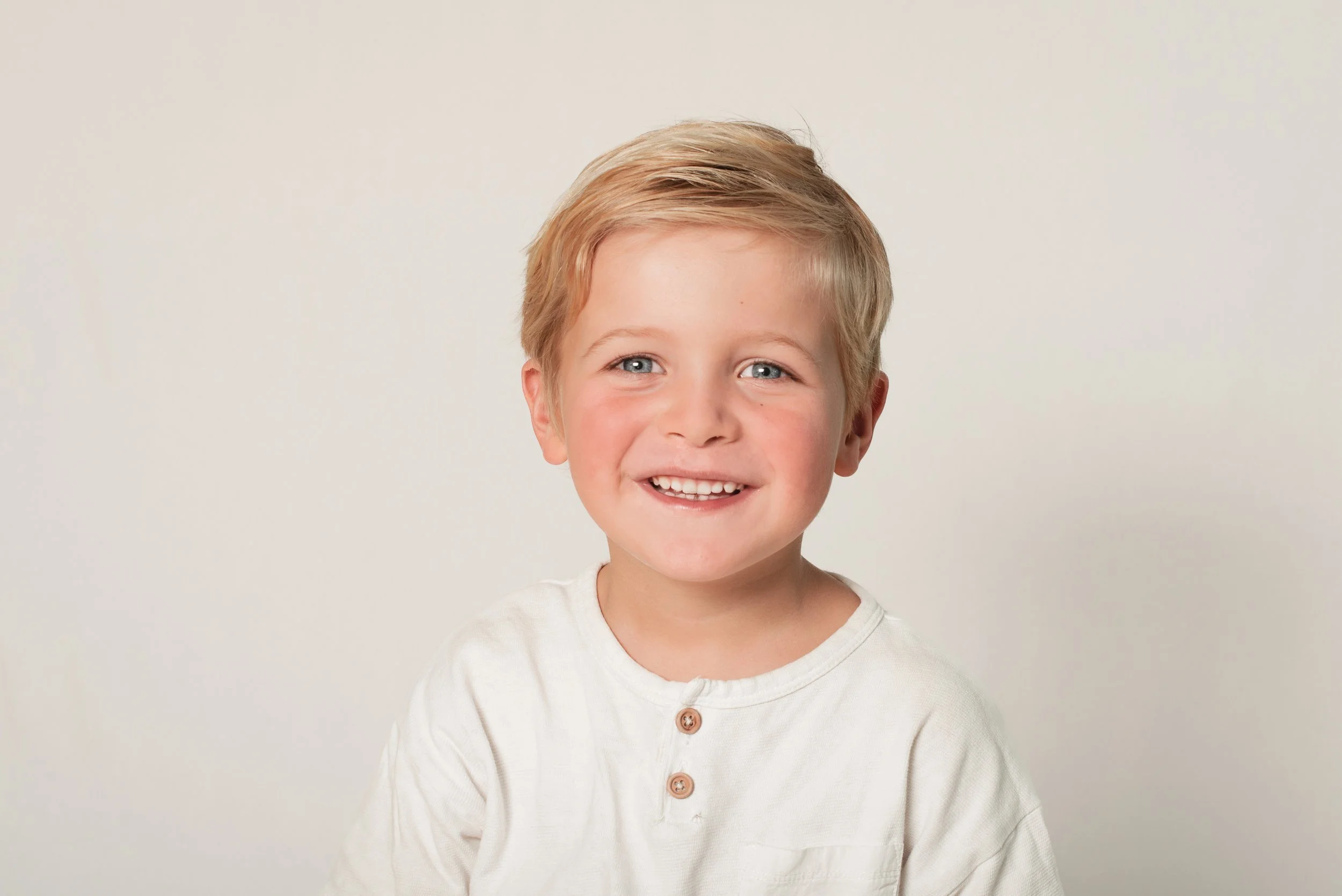 A young boy with short blonde hair, blue eyes, and a cheerful smile, wearing a white shirt with brown buttons, standing against a plain light-colored background.