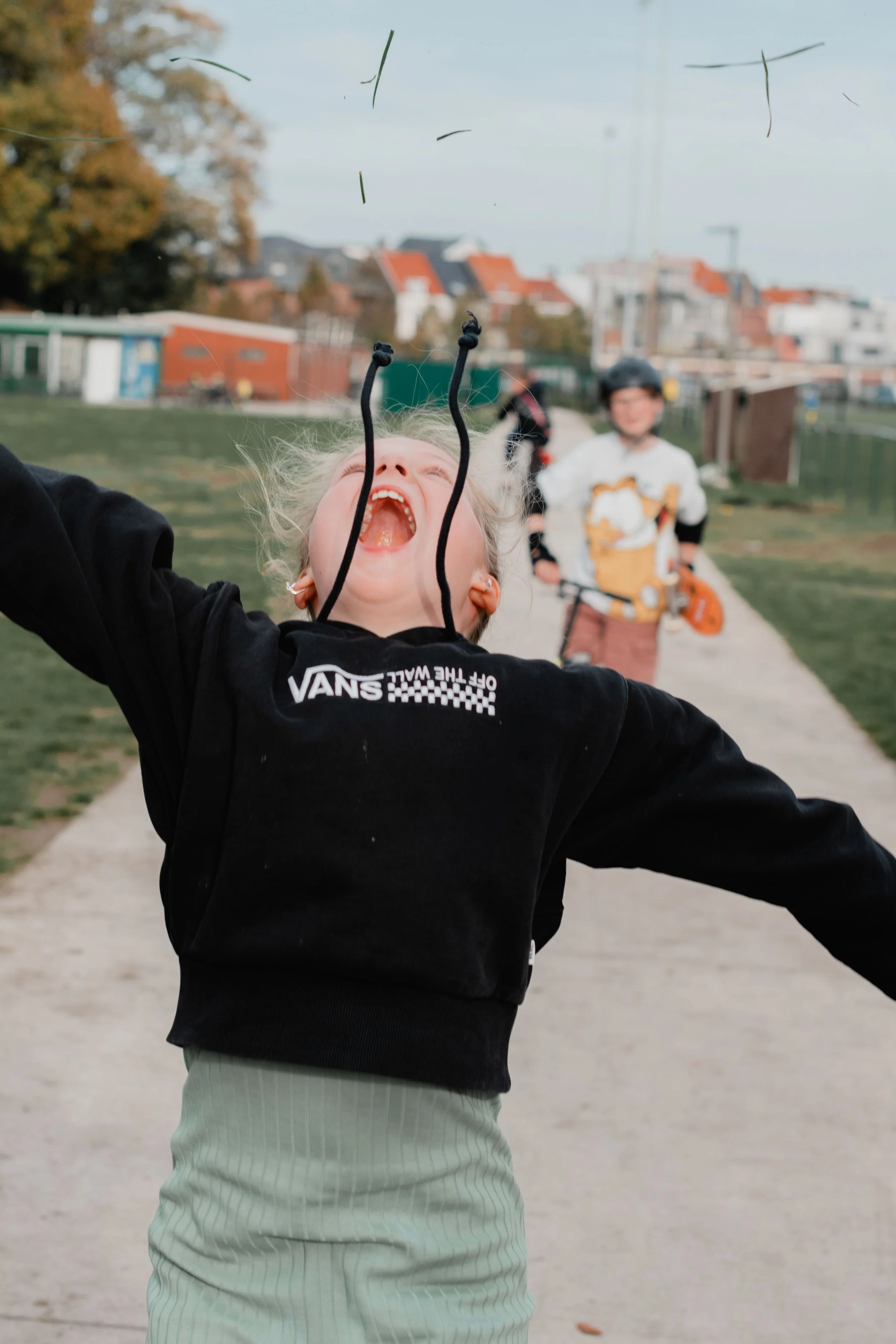A young person with blond hair and earrings is joyfully throwing grass or small plants into the air outdoors, with a person on a bicycle and a residential neighborhood with houses in the background.