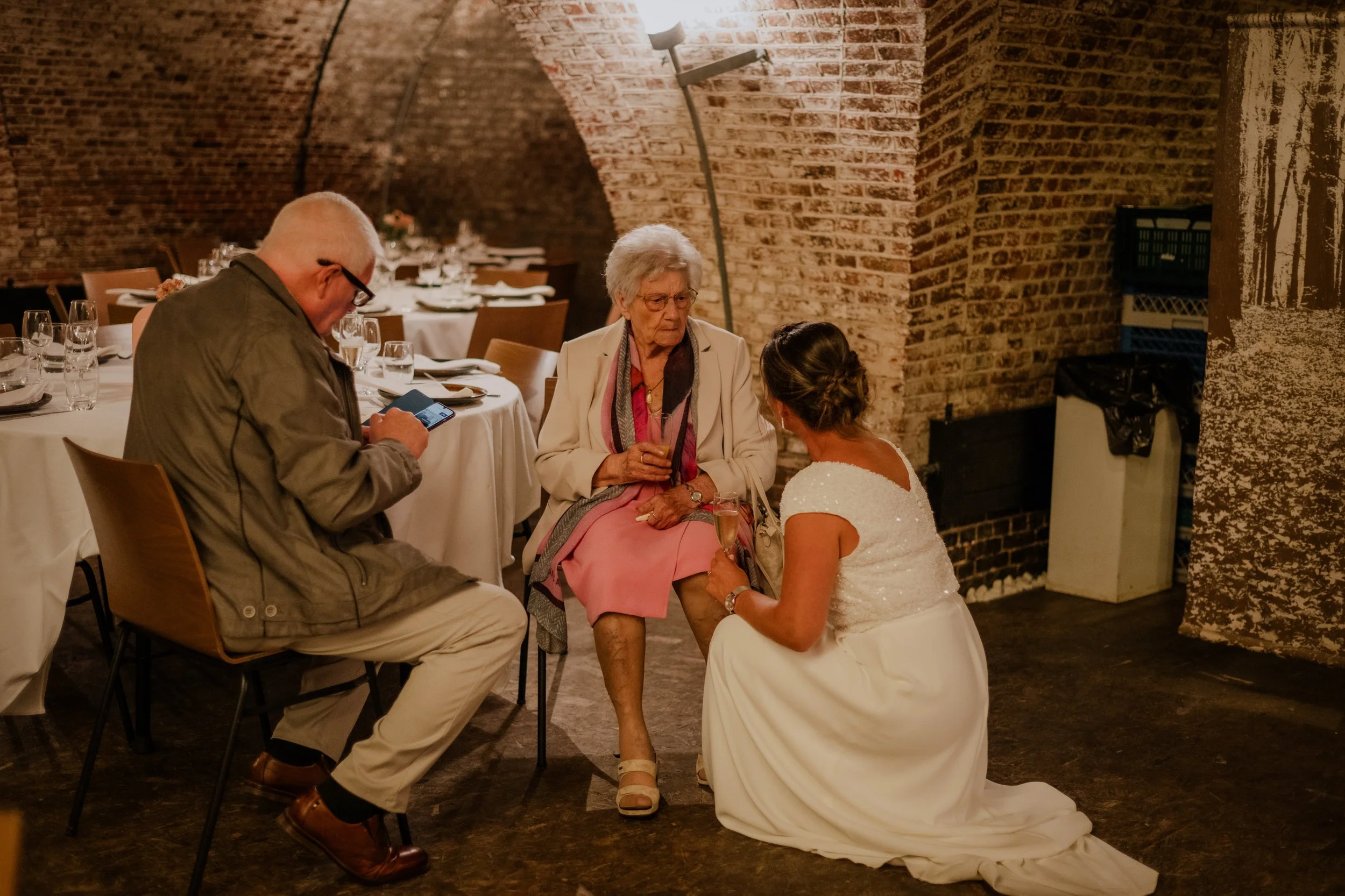 An elderly woman, dressed elegantly, is sitting in a restaurant with a younger woman in a wedding dress kneeling in front of her, holding her hand. A man seated nearby is looking at his phone. The restaurant has brick walls and formal table settings.