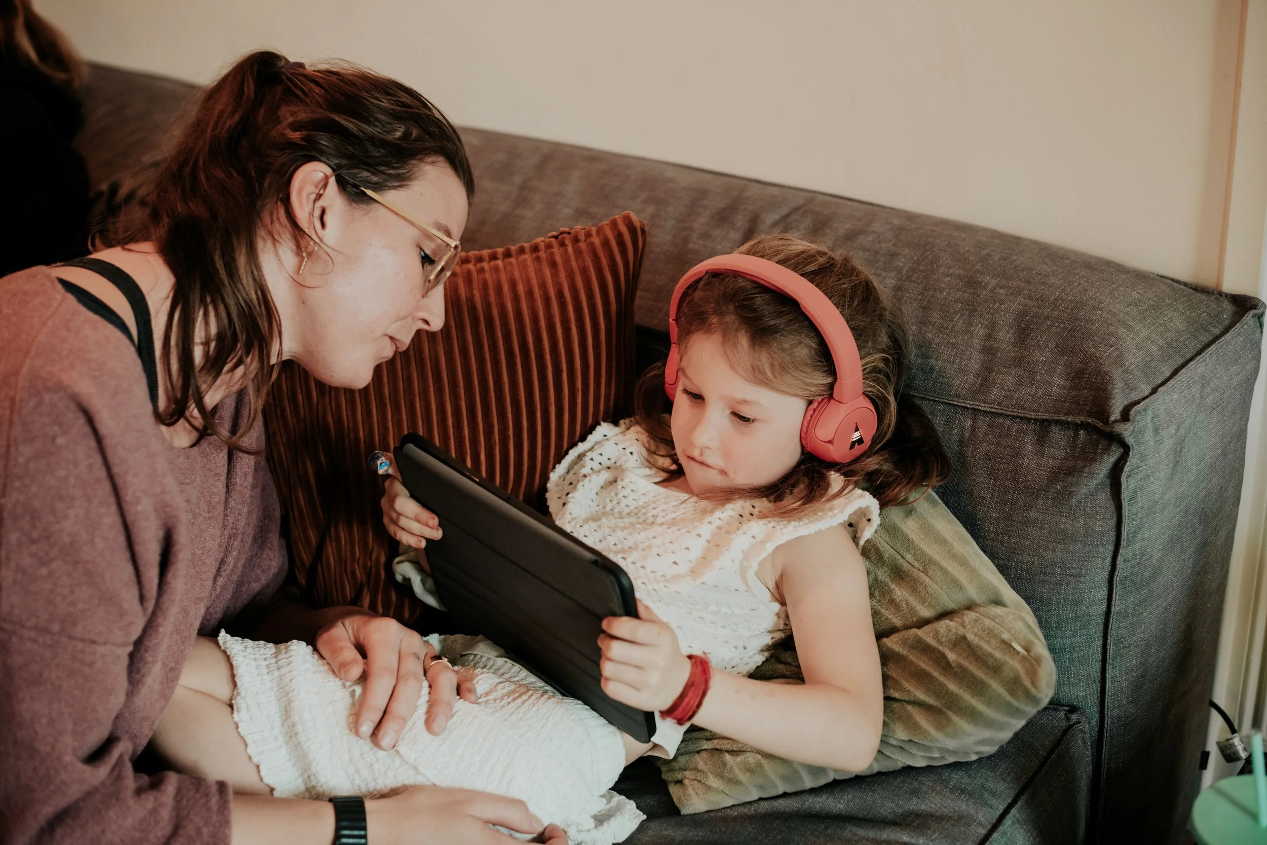 A woman with glasses and a young girl with headphones looking at a tablet together while sitting on a couch.