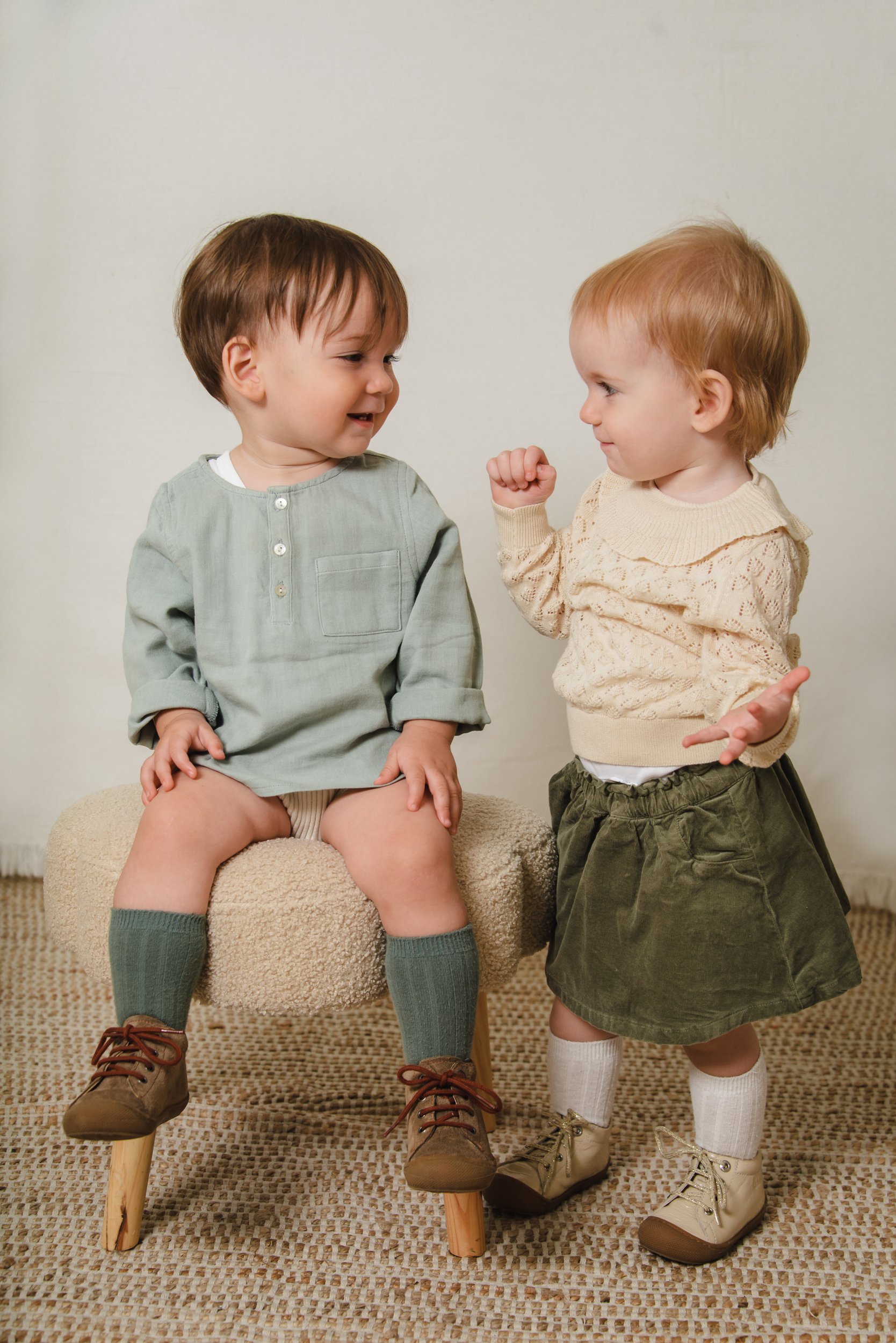 Two young children, a boy and a girl, face each other and smile. The boy sits on a beige stool with chunky brown shoes, green socks, and a light green shirt. The girl stands, leaning slightly forward, wearing beige boots, white socks, and a cream-col