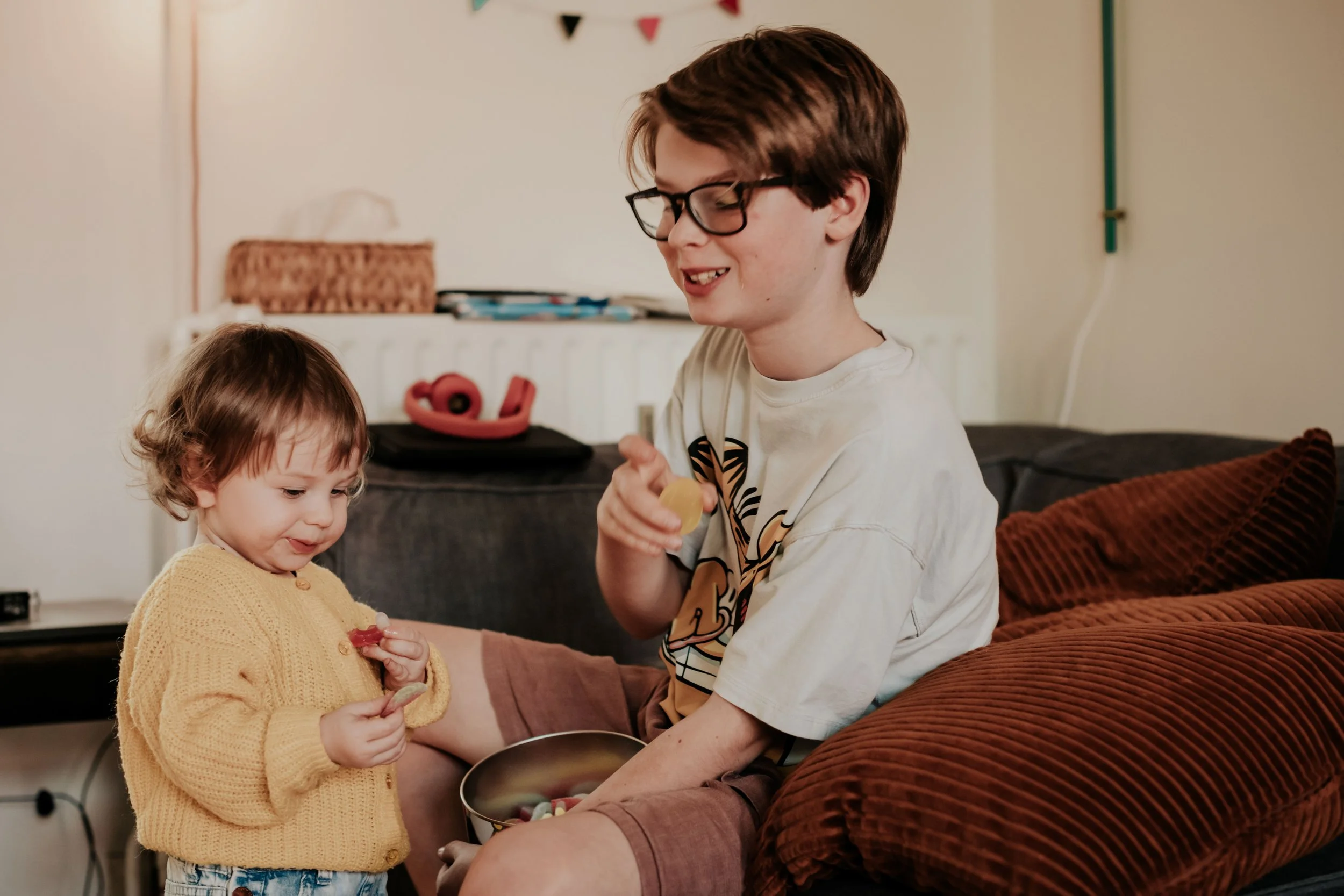 Two children, a young girl with short curly hair wearing a yellow sweater and a teenage boy with glasses and brown hair, are sitting on a couch. The girl is holding a snack, and the boy is holding a bowl filled with candy. They are in a cozy, decorat