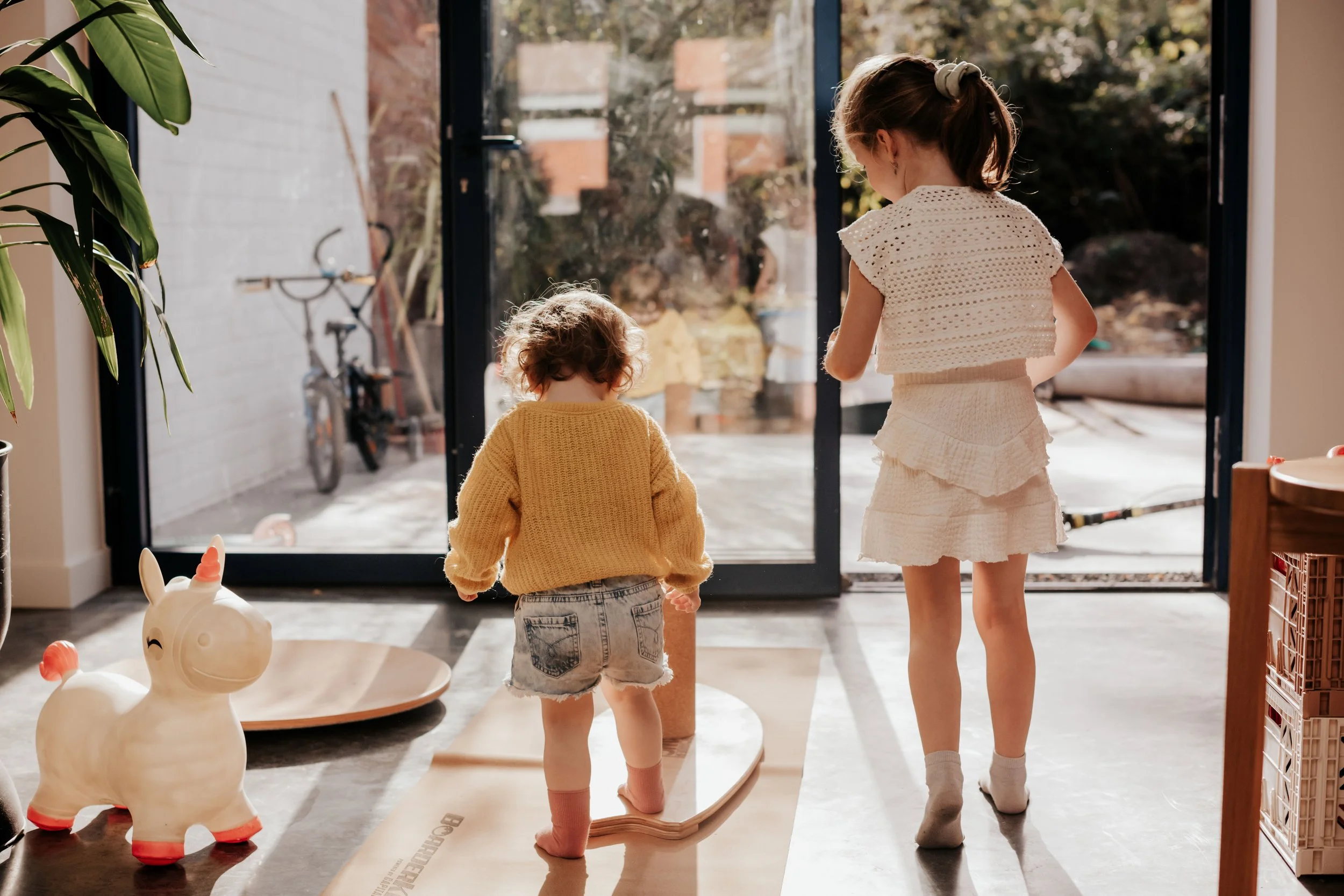 Two young children, a girl and a boy, are standing indoors near a large glass door, looking outside. The girl has long hair tied back, wearing a white skirt and a crocheted top. The boy has curly hair, wearing a yellow sweater and denim shorts. There