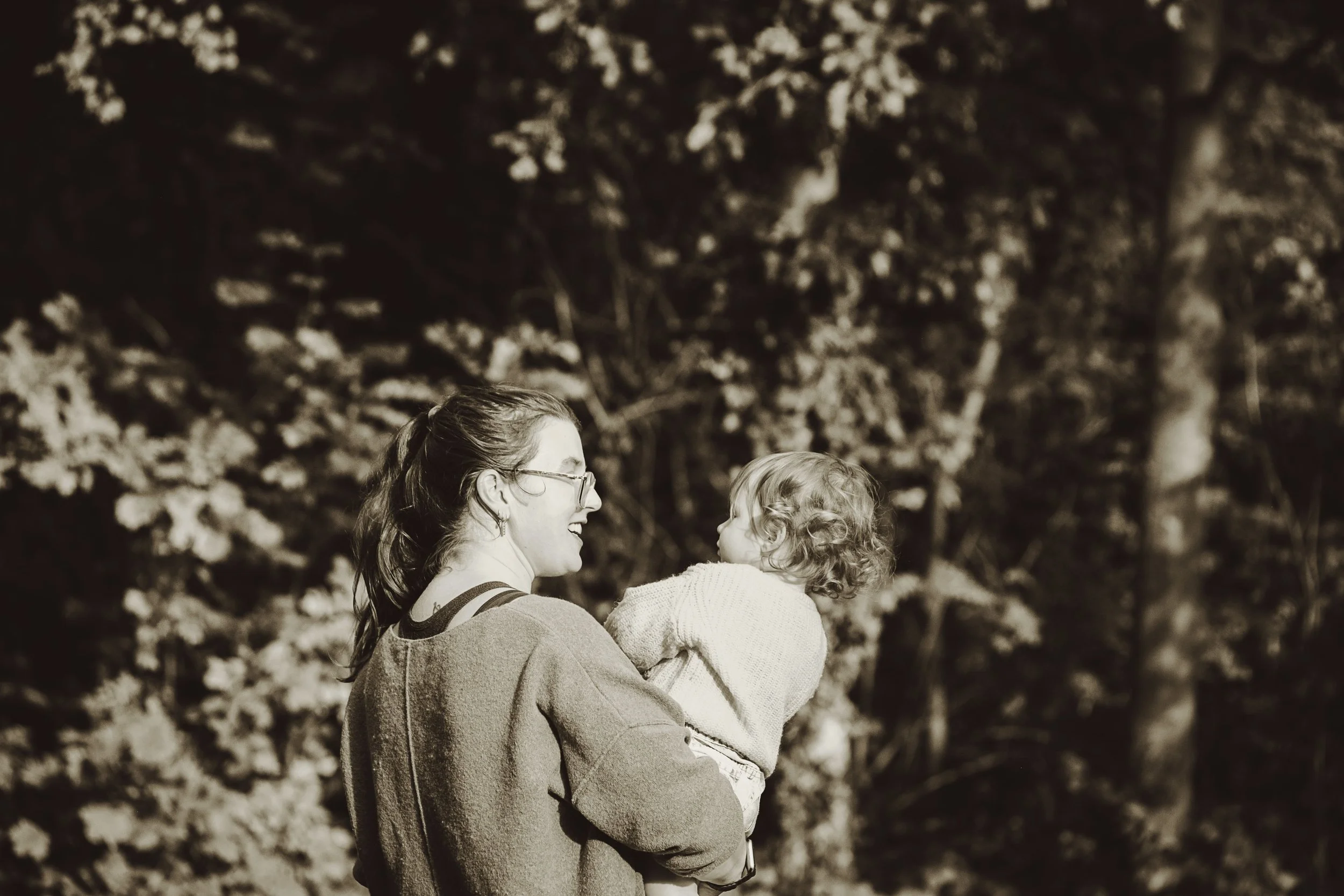 A woman with glasses holding a young girl, who is wearing a sweater, outdoors with trees in the background.