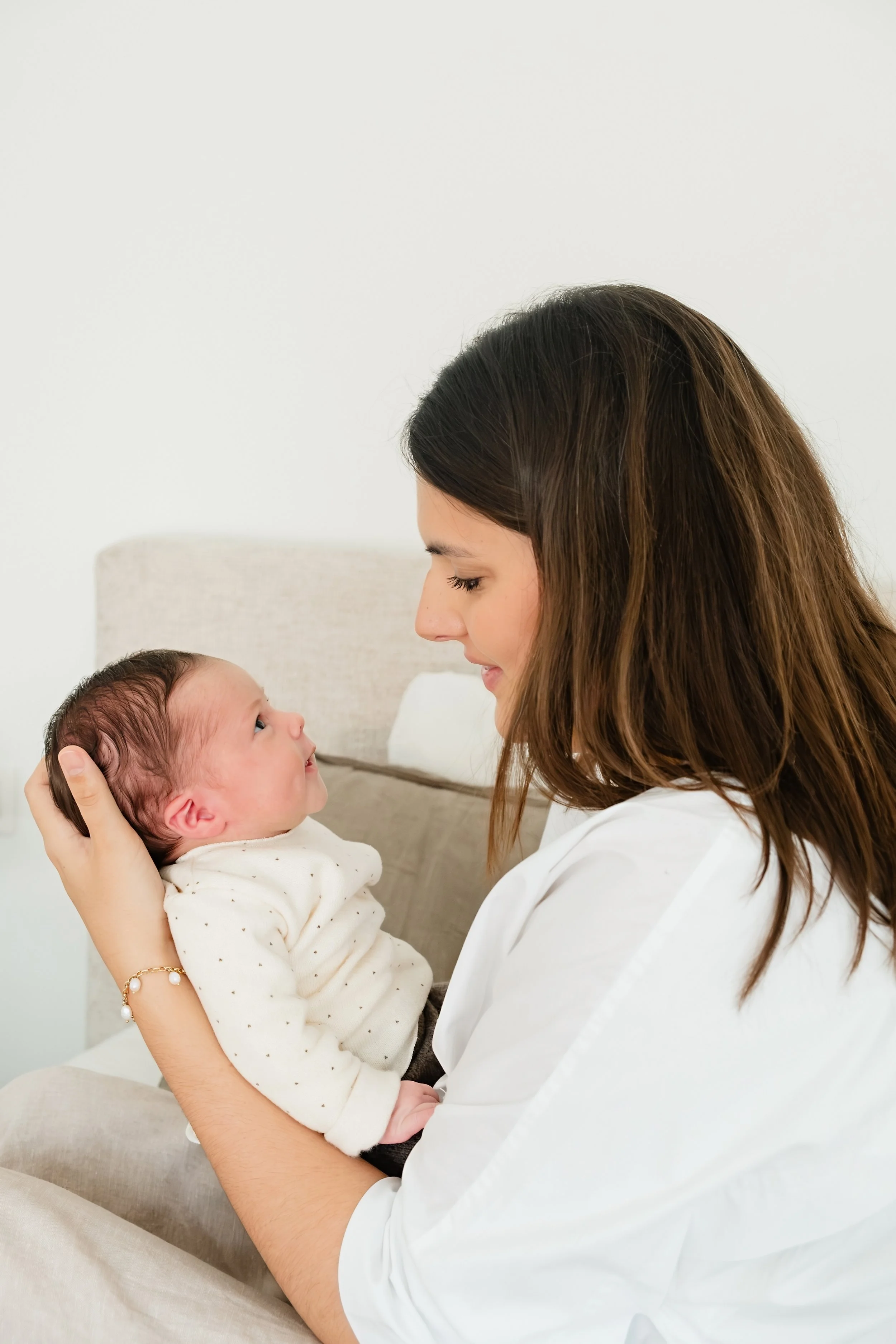 A woman holding a newborn baby in her arms, both looking at each other lovingly. The woman has long brown hair and is wearing a white shirt. The baby is dressed in a white and tan polka-dotted onesie, resting comfortably in the woman's hands.
