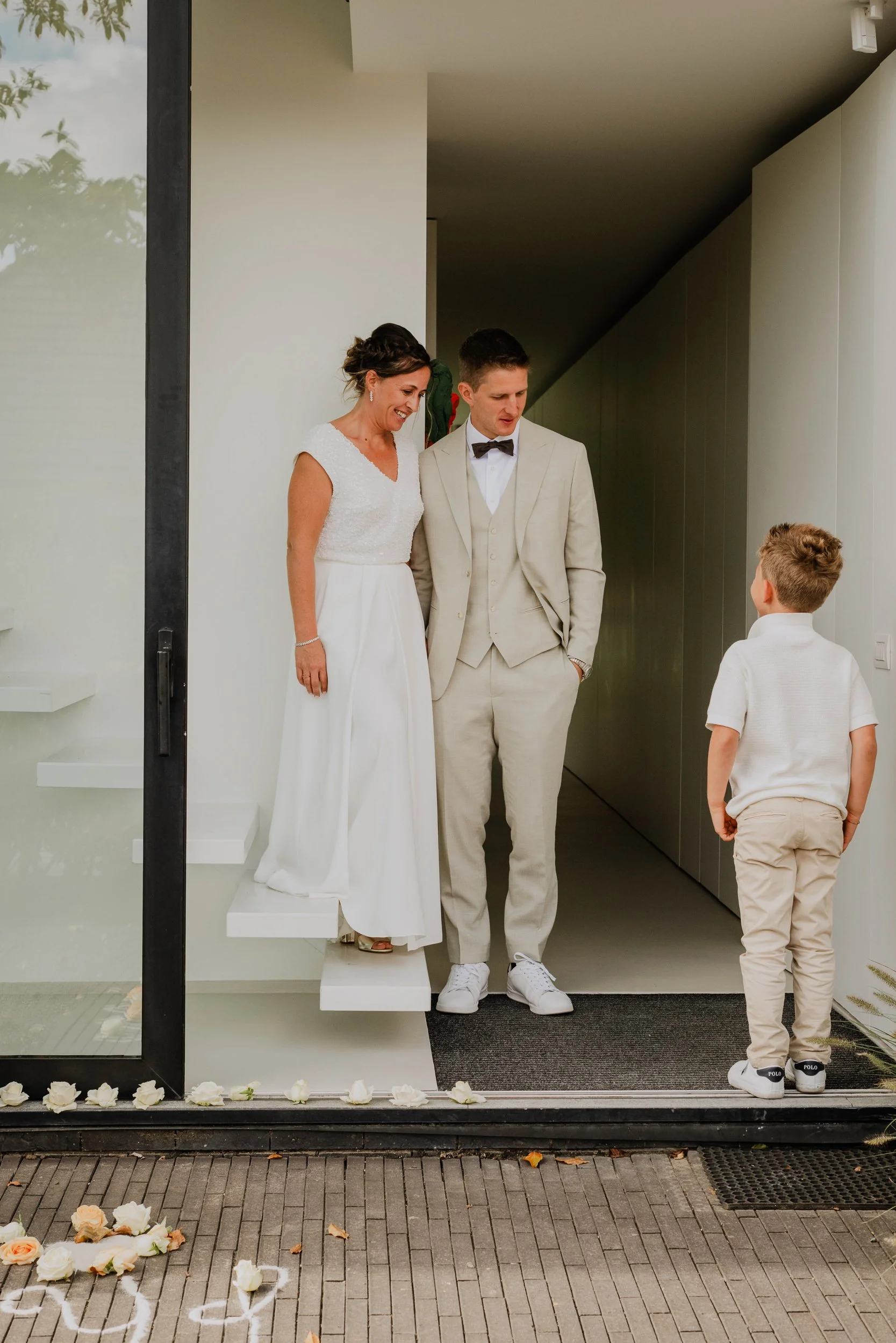 A bride and groom standing under an awning inside a modern building, looking at a young boy facing them. The scene is decorated for a wedding, with white roses along the floor and on a small staircase.