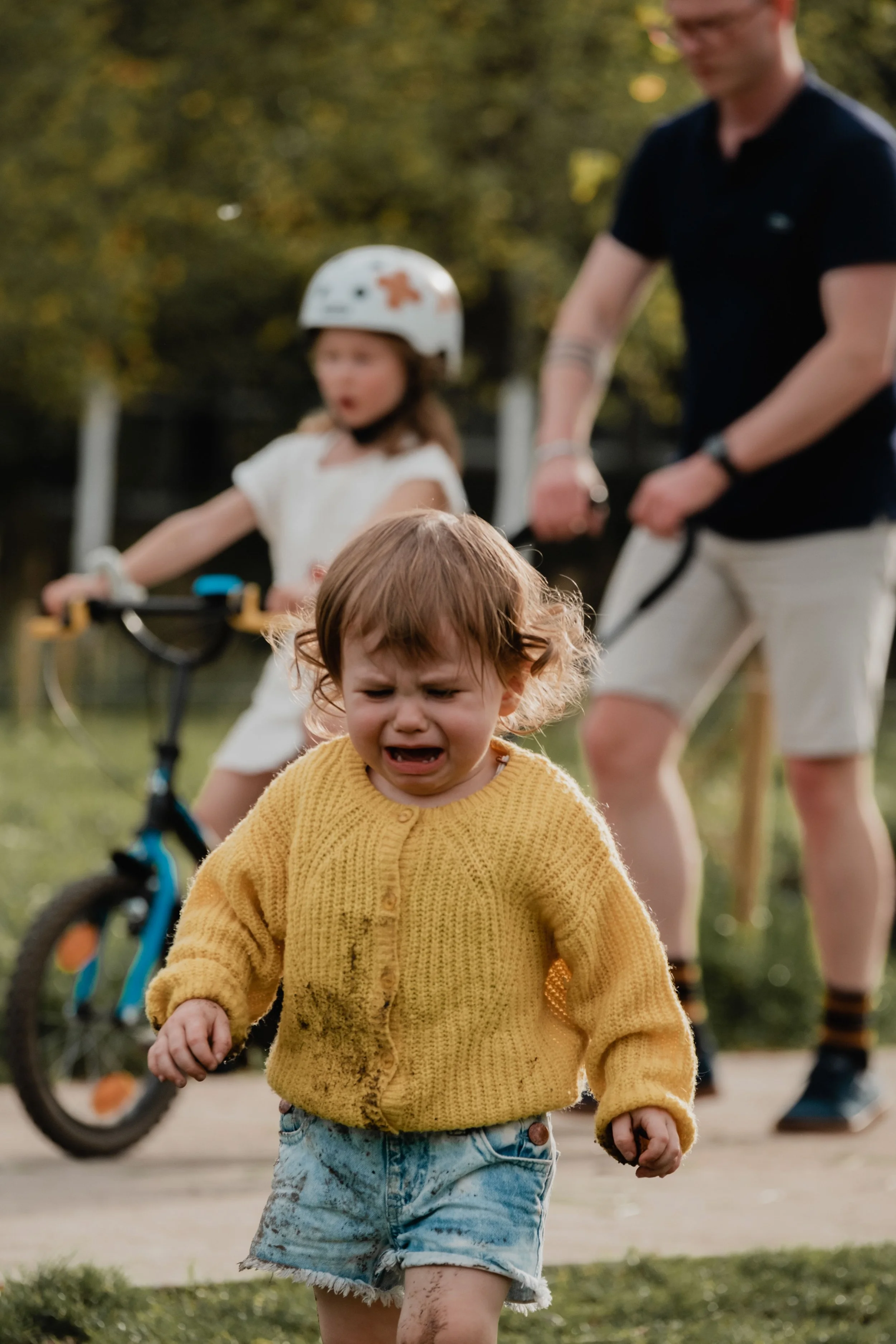 Child wearing a yellow sweater and denim shorts crying while running outdoors, with a woman walking a bike and a girl in a helmet behind, in a park or backyard setting.