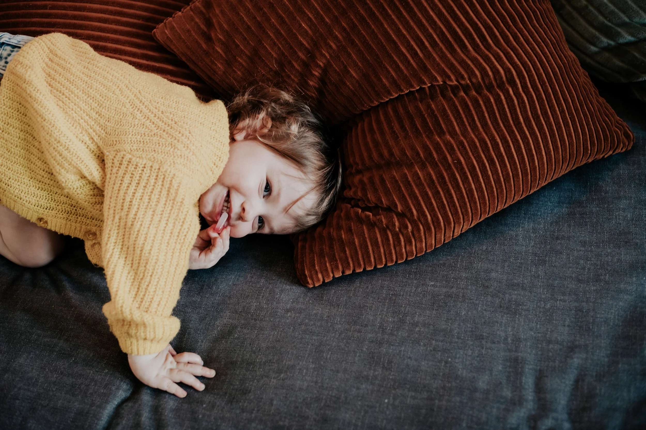 A young child lying on a dark sofa, partially covered with a yellow knitted blanket, holding a toothbrush, smiling with blue eyes, surrounded by colorful pillows.
