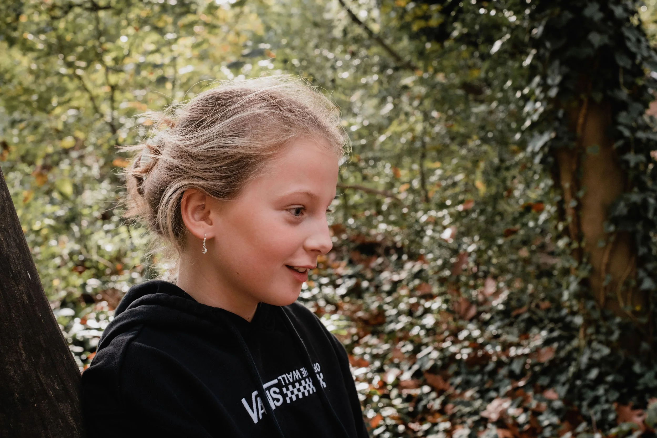Young woman with blonde hair wearing a black hoodie and earrings, standing outdoors near a tree with green and orange leaves.