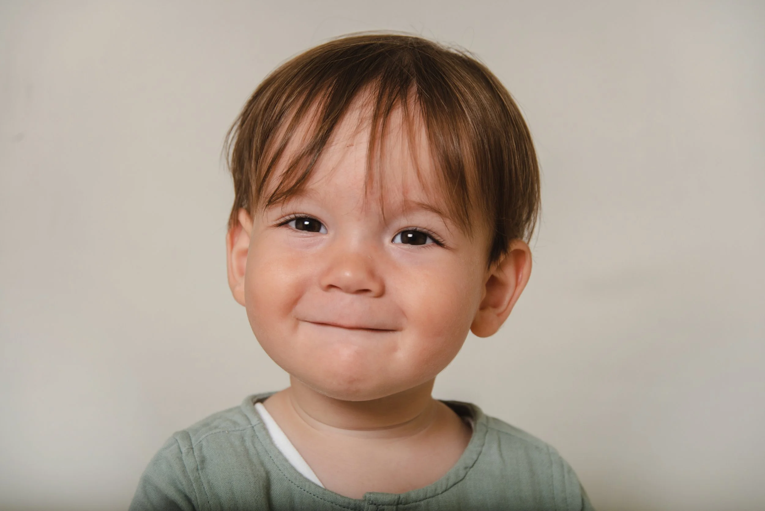 Close-up of a smiling young boy with short brown hair, brown eyes, and wearing a green shirt with a white collar, against a plain light background.