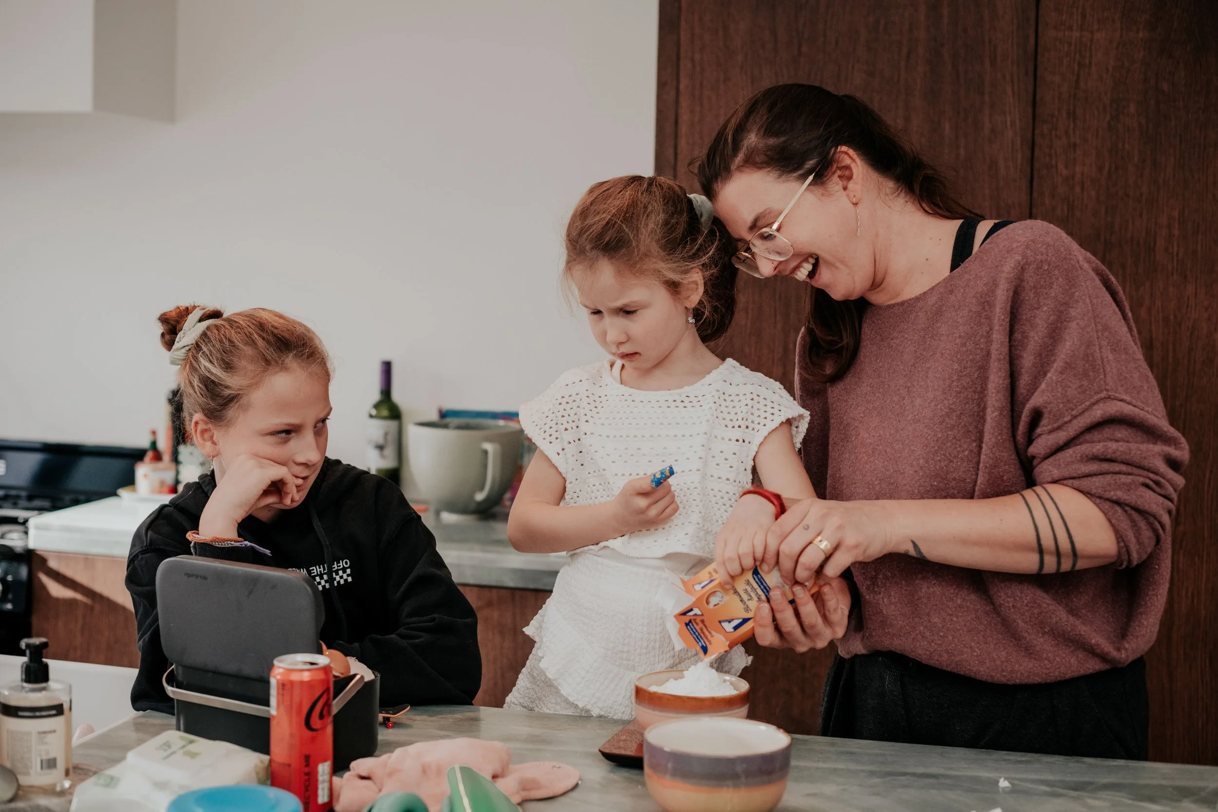 A woman with glasses smiles while interacting with two young girls in a kitchen or classroom setting. One girl has her hand near her face, and the other girl is holding a snack or small object.
