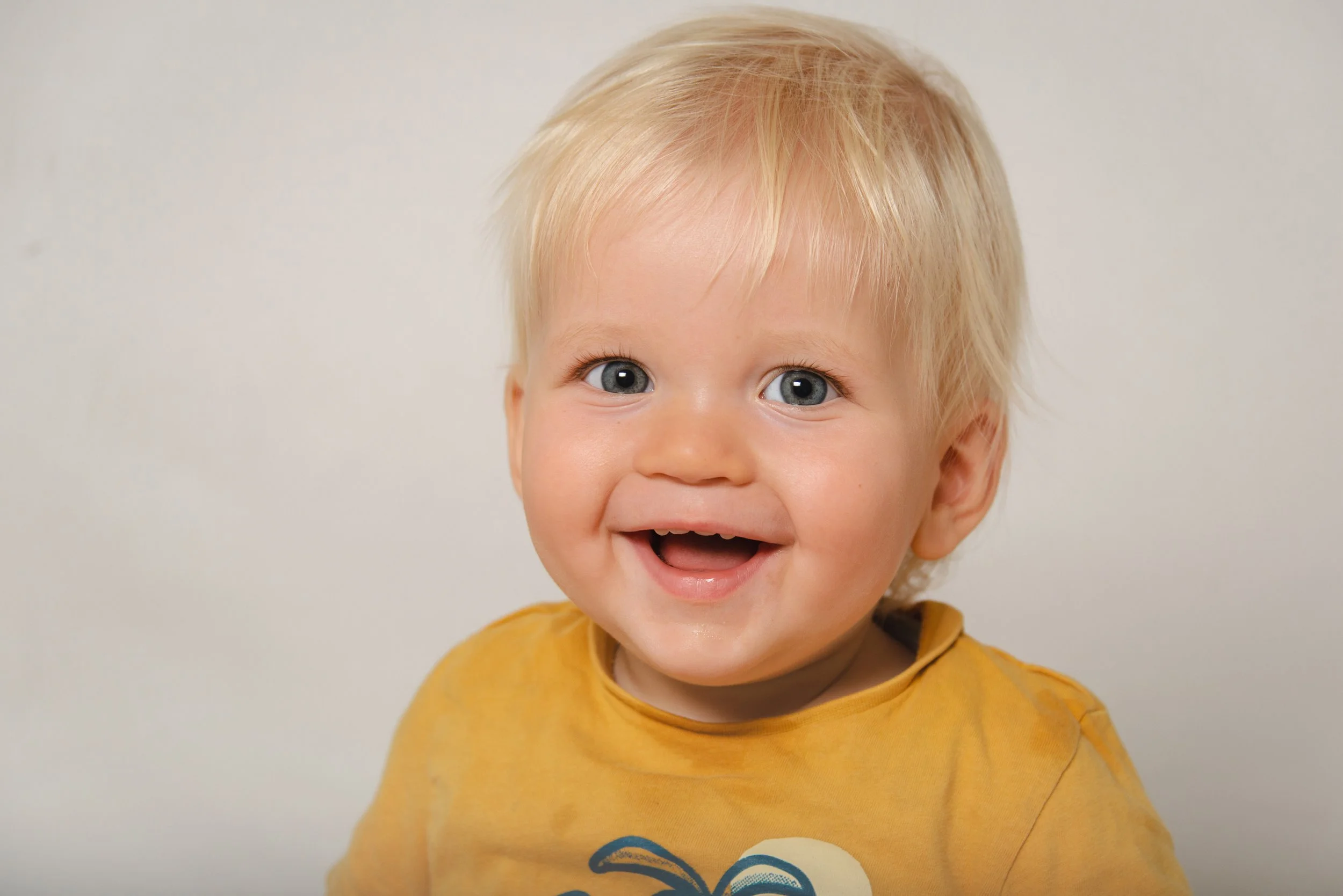 A young child with blonde hair, blue eyes, and a big smile, wearing a yellow shirt with a blue design, against a plain white background.