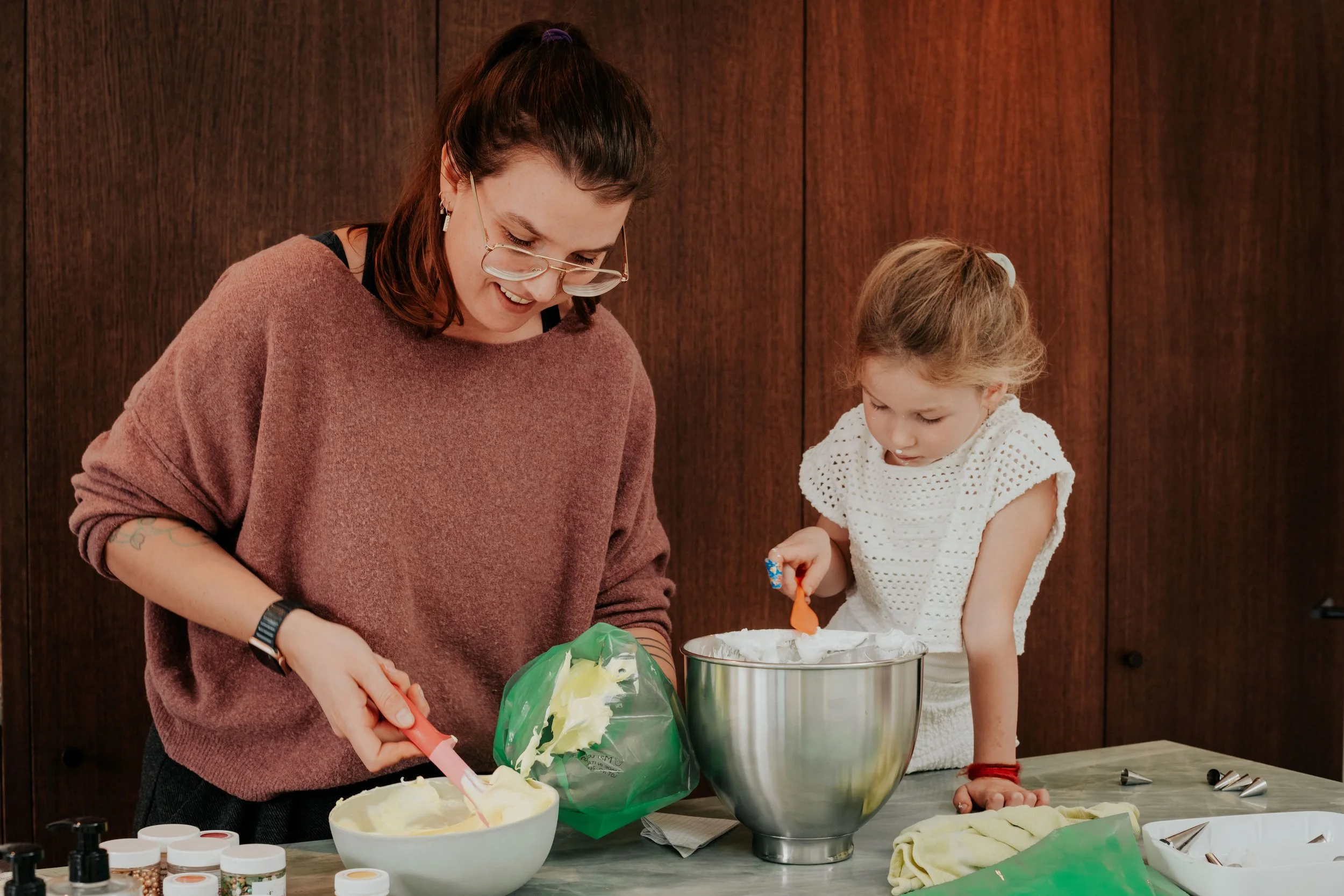 A woman and a young girl are decorating a cake together in a kitchen or bakery. The woman has brown hair, glasses, and is wearing a pinkish sweater. The girl is wearing a white dress with eyelet details and has light brown hair tied back with a white