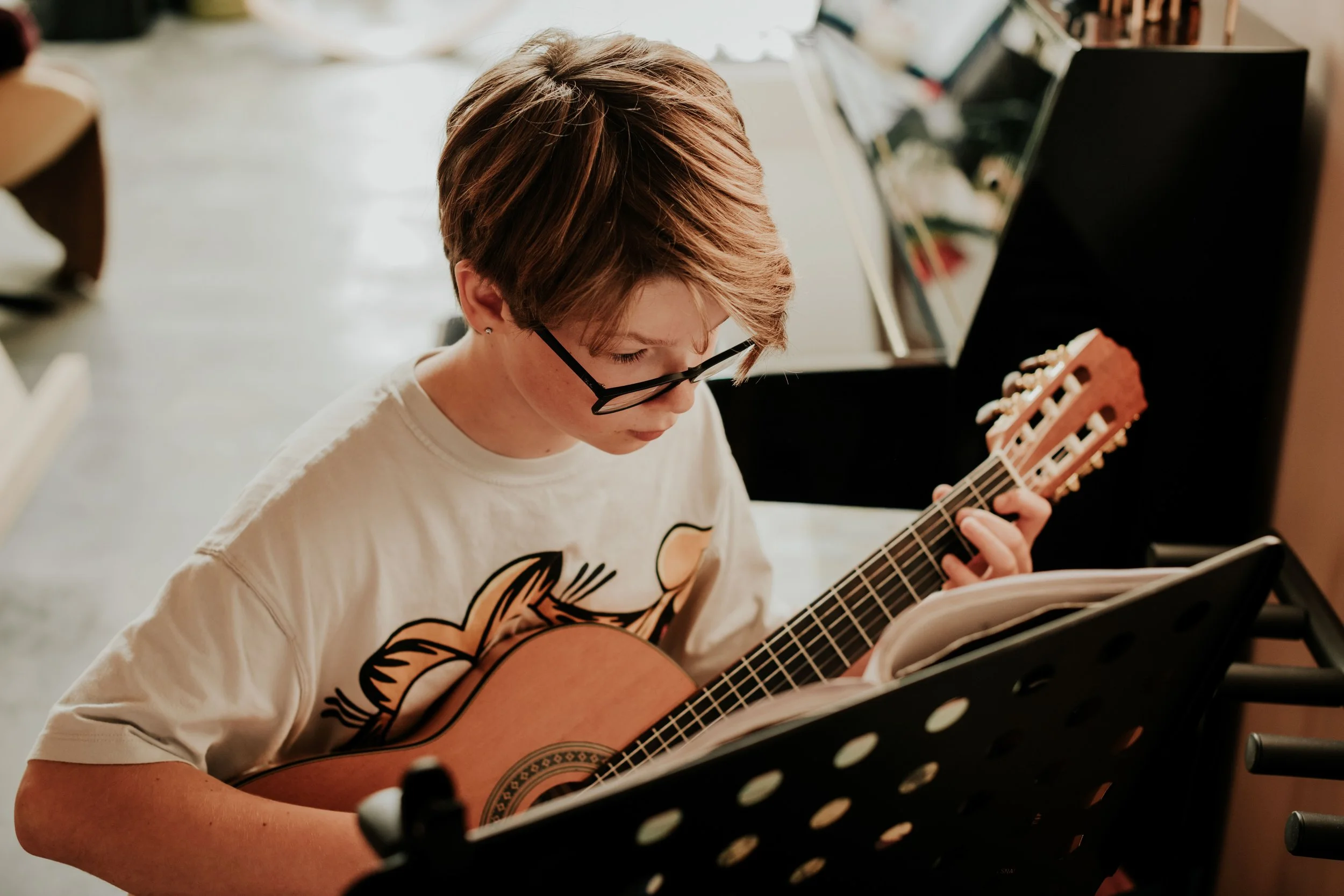 A young person with short hair and glasses playing an acoustic guitar, sitting in front of a music stand, in a room with a piano and other furniture.