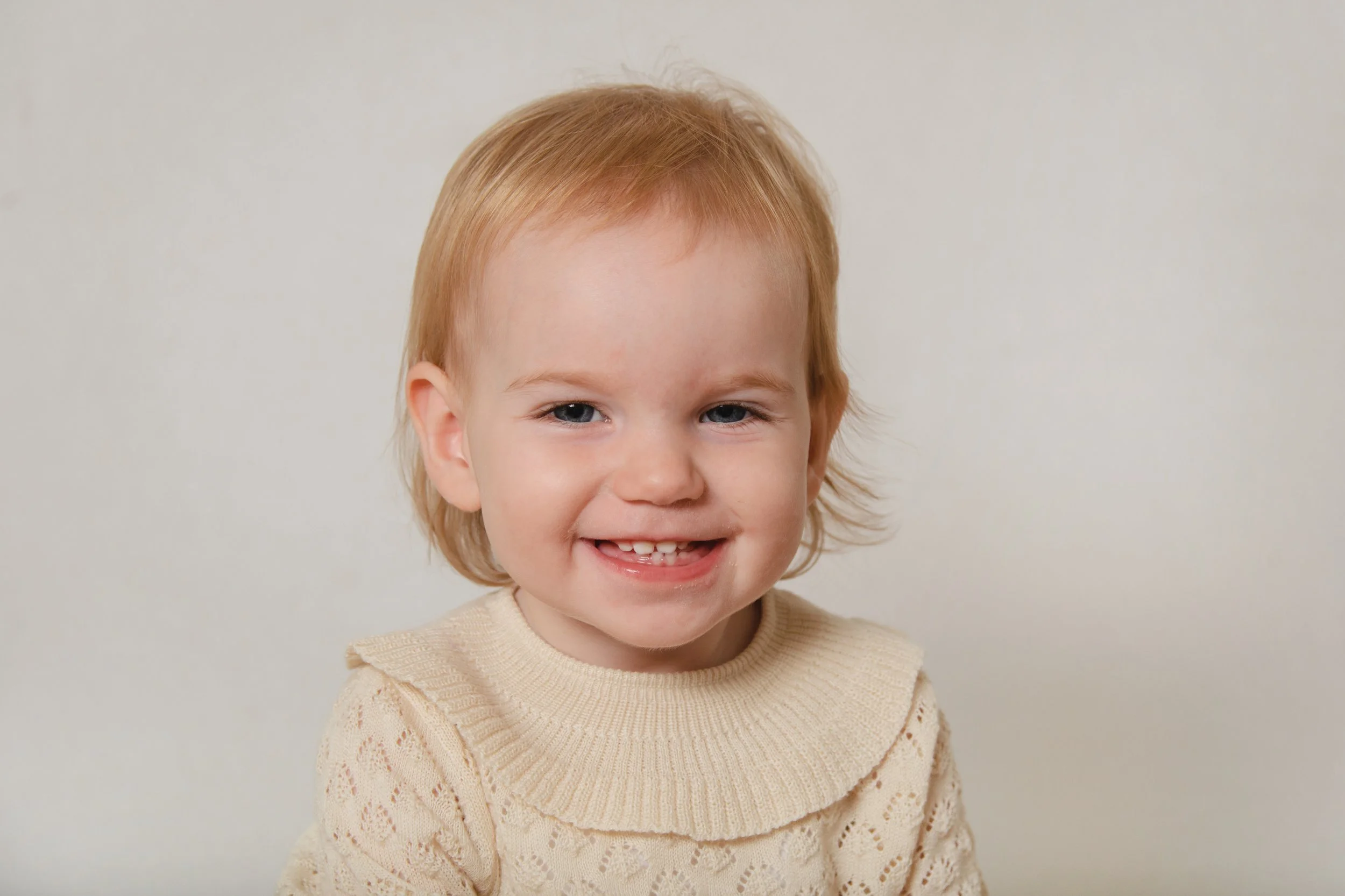 A young child with red hair and blue eyes smiling, wearing a beige sweater, standing against a plain light-colored wall.