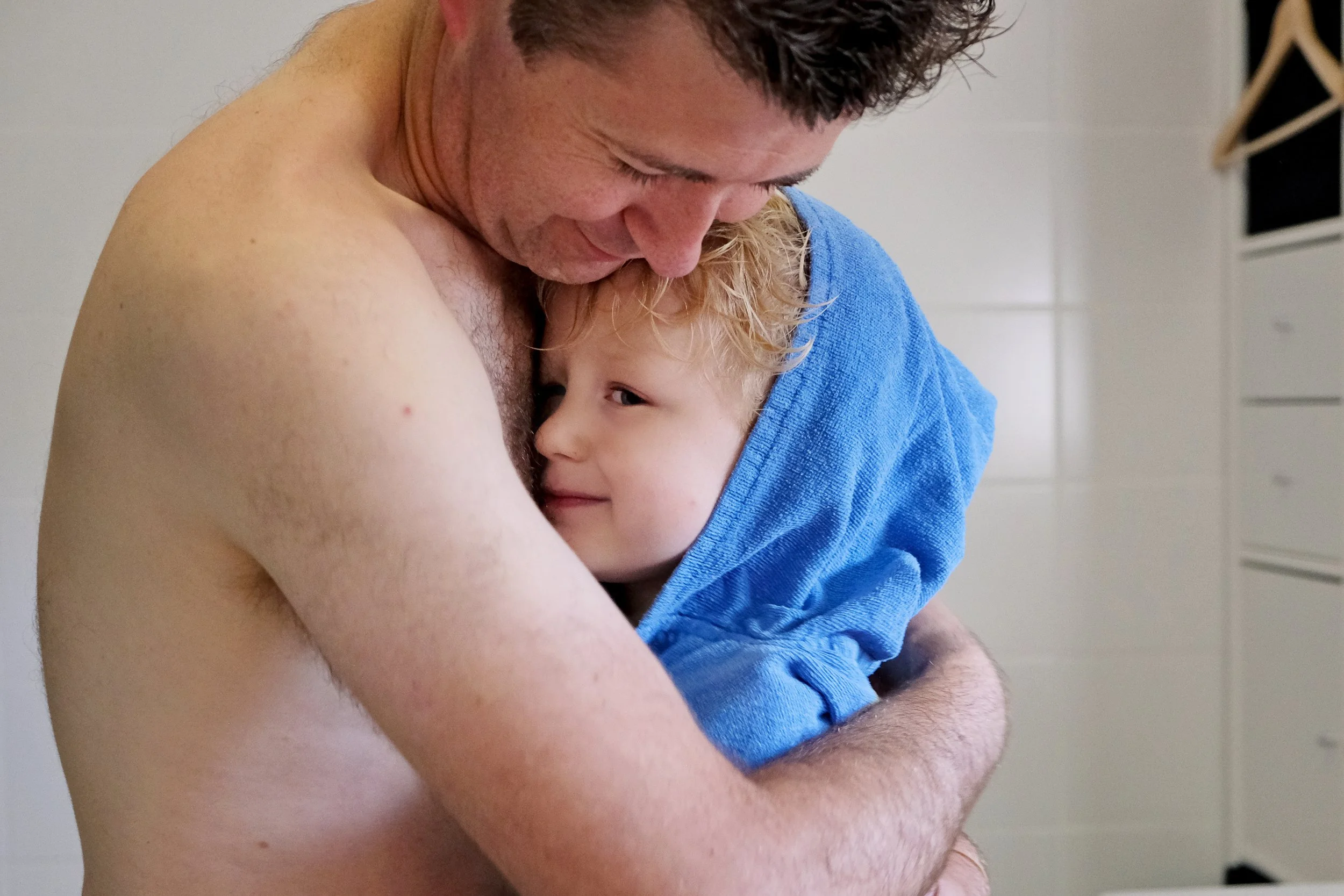 A man embraces a young boy wrapped in a blue towel, with both appearing to be happy and content. The man has short dark hair and is looking down, while the boy has curly blonde hair and is smiling.