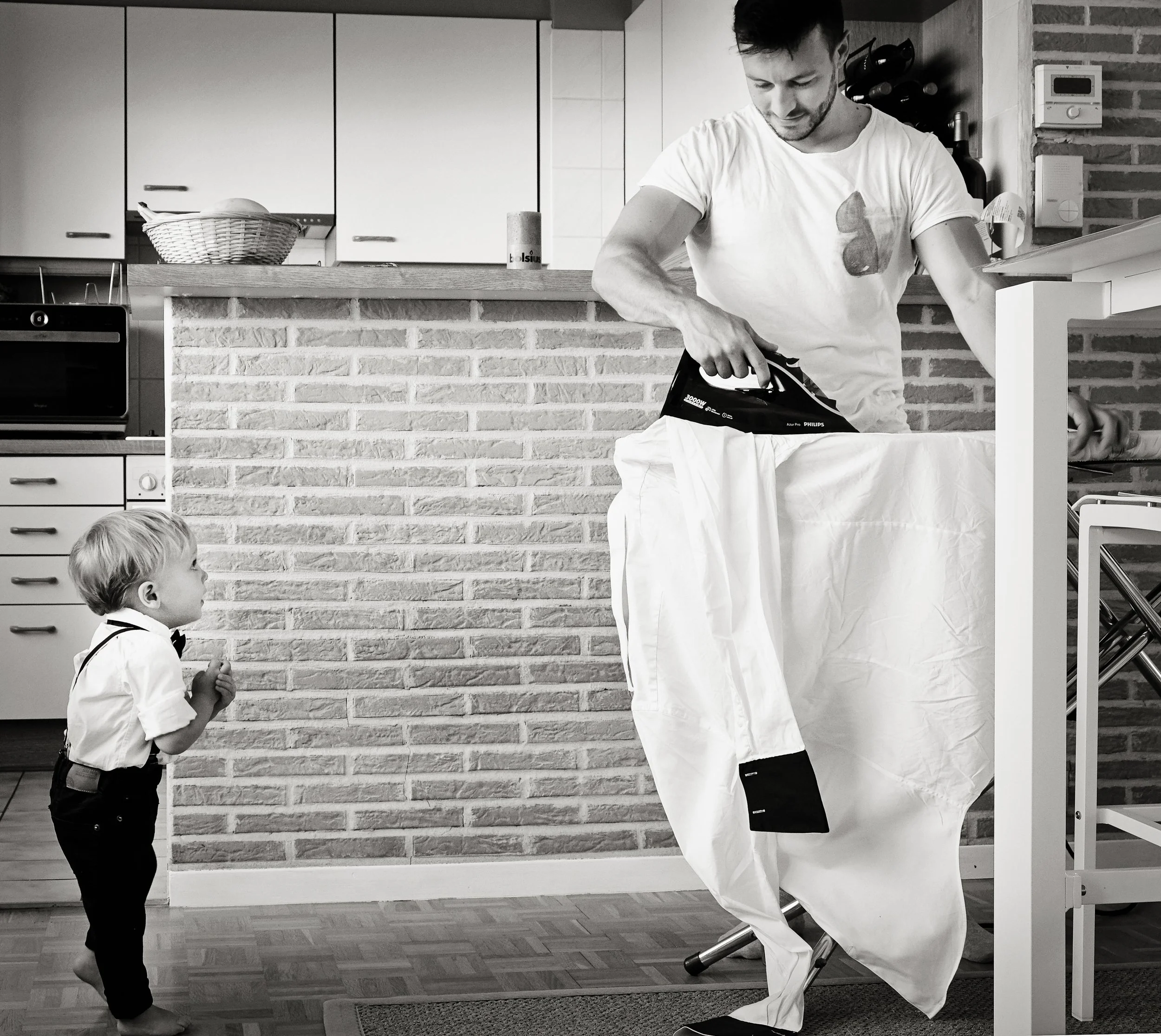 A young man and a young boy in a kitchen, with the man ironing clothes and the boy watching. The man is wearing a white t-shirt and white pants, while the boy is dressed in a white shirt with a bow tie.