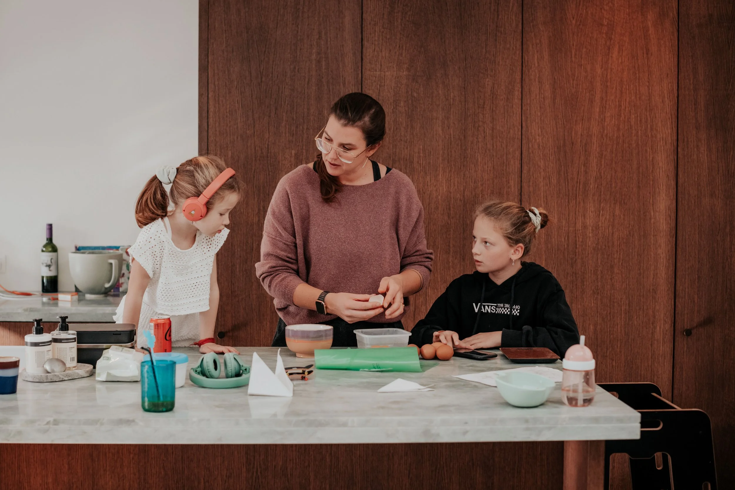 A woman with glasses and dark hair in a ponytail is talking to two young girls at a kitchen counter. One girl has blonde hair, wearing pink headphones and a white shirt, and the other girl has light brown hair tied back and is wearing a black hoodie.