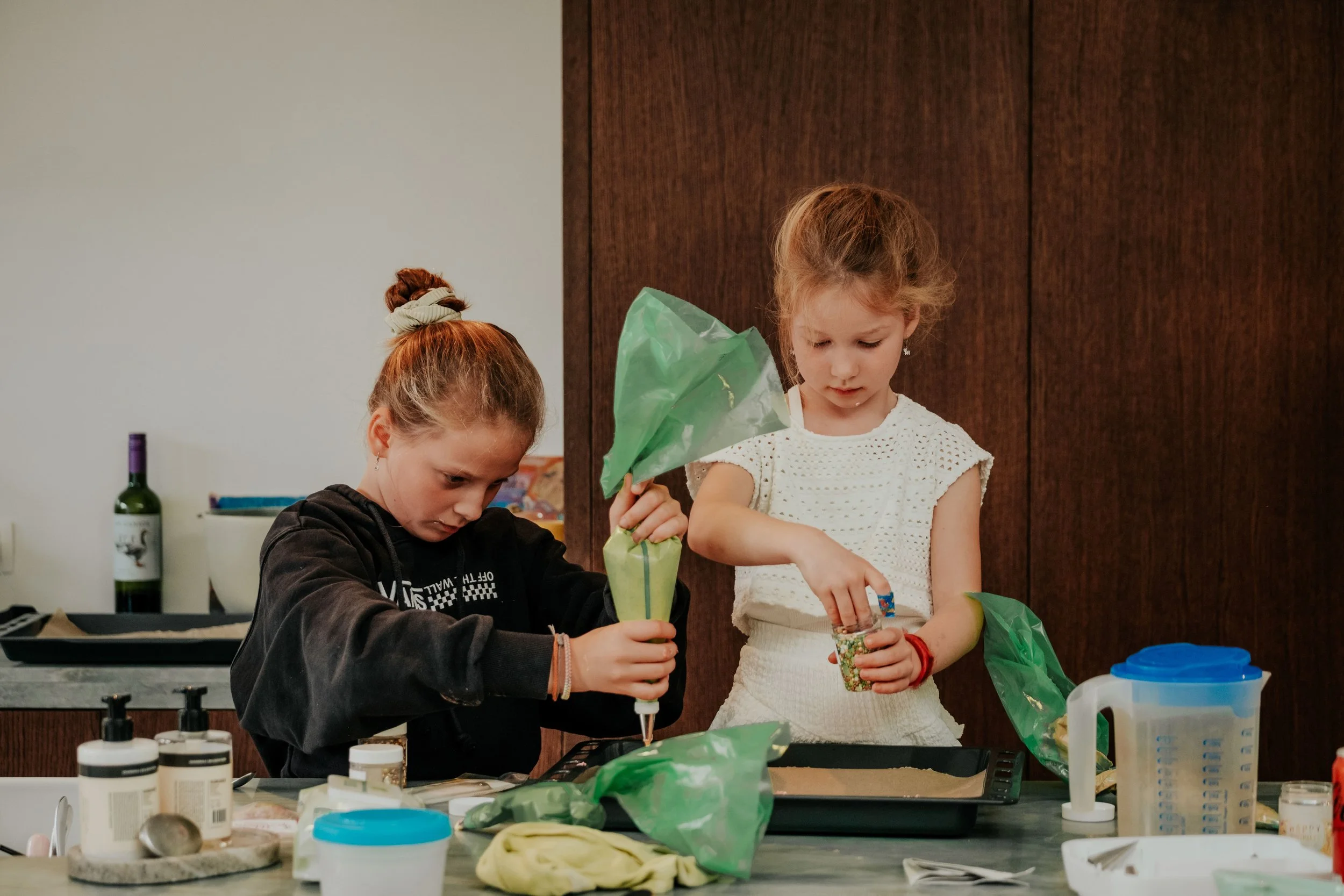 Two young girls working on a craft project at a table with various supplies, including bottles, bowls, and bags, in a room with a wooden wall.