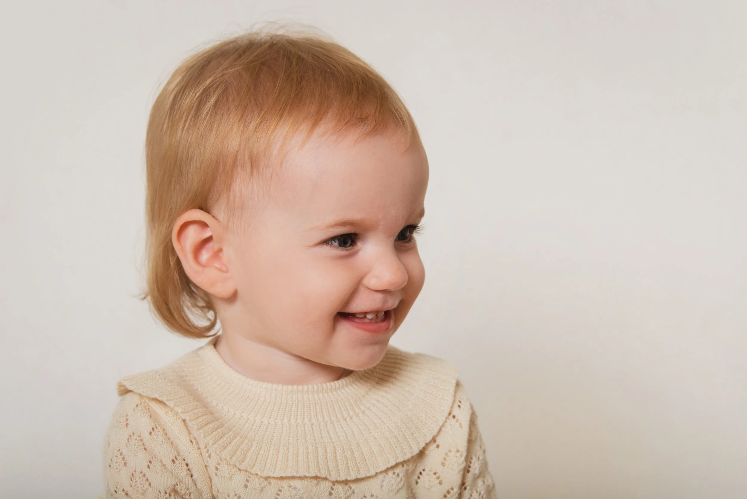 A young girl with red hair smiling and looking to her right, wearing a beige sweater with a ruffled collar, against a plain white background.