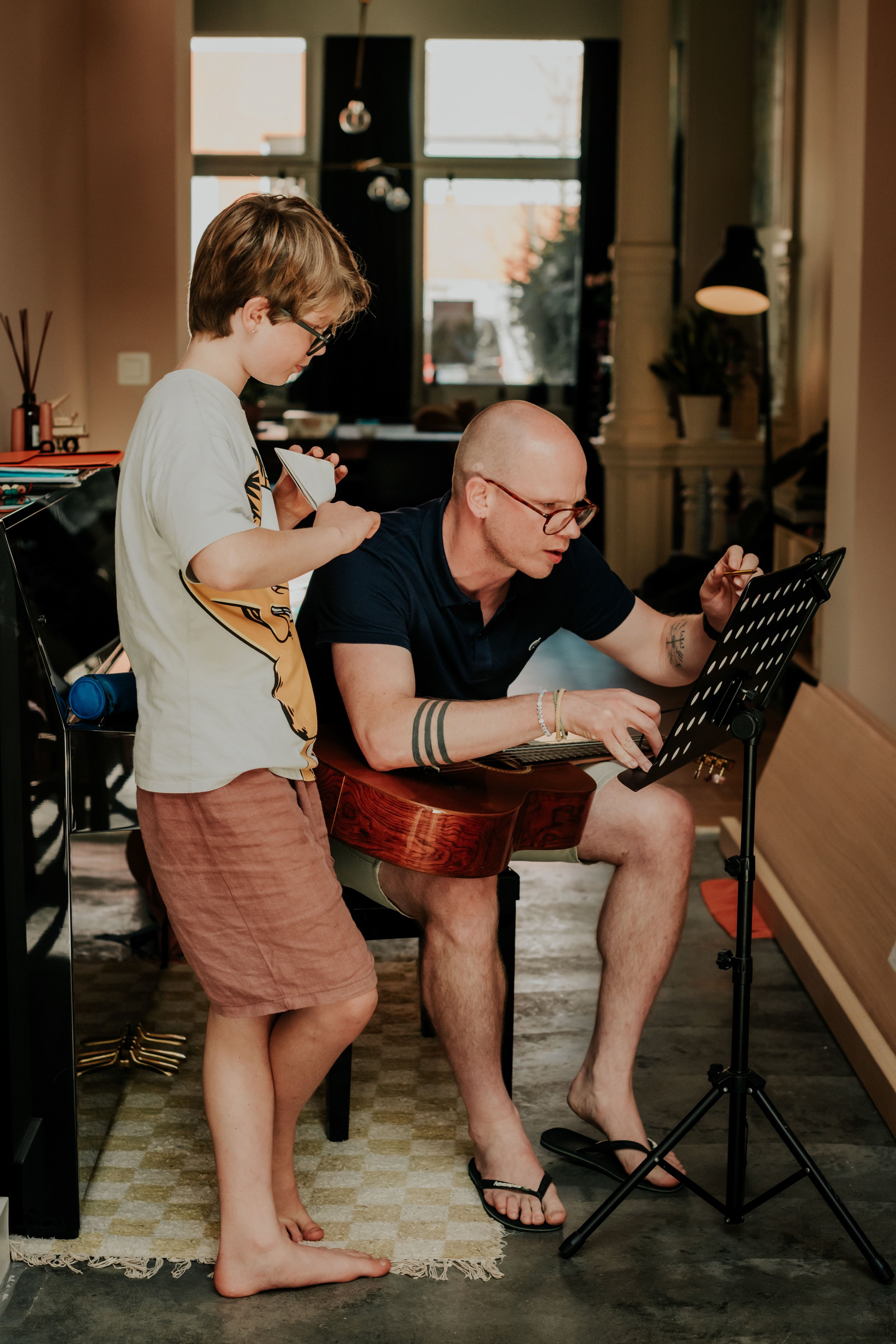 A young boy and an older man are in a room. The boy is standing, holding a small notepad, while the man is sitting at a desk, reading music sheets with a music stand in front of him. The boy is barefoot, and the man is wearing flip-flops. The backgro