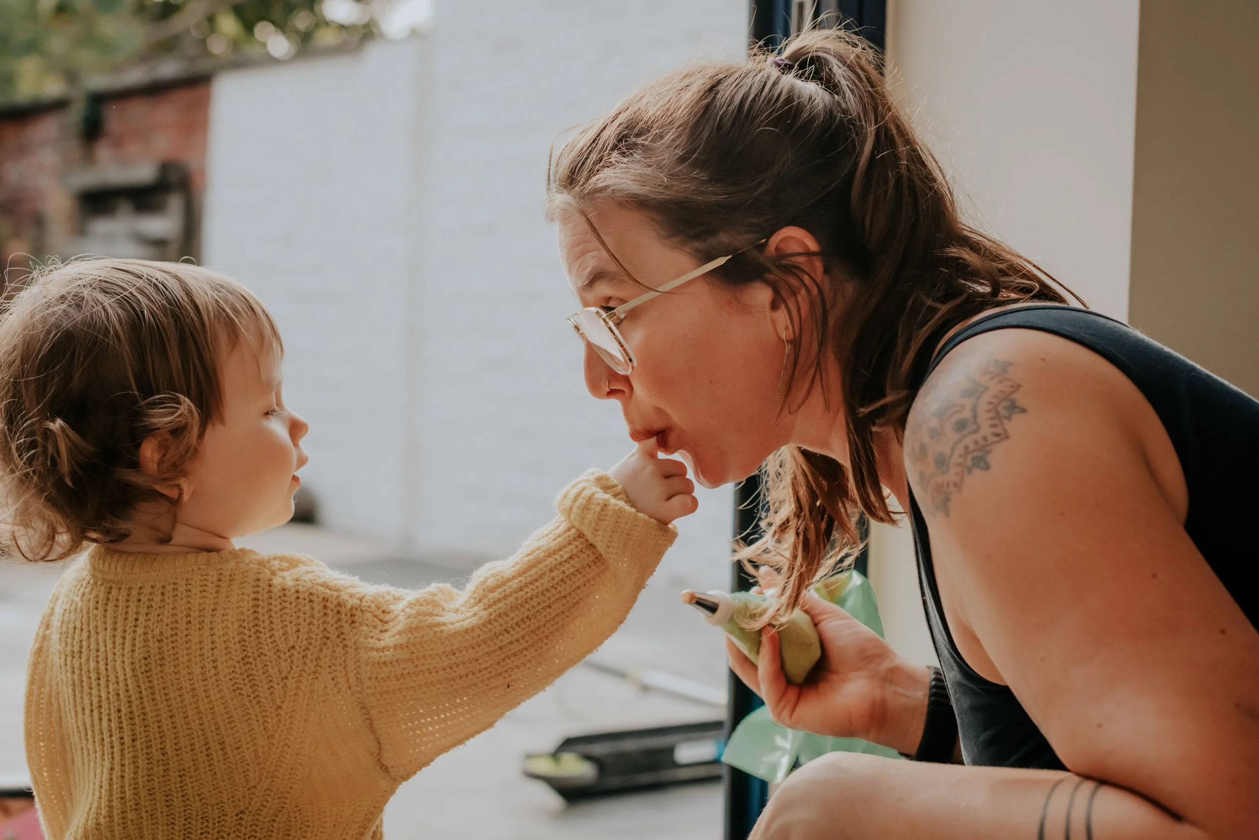 A young girl in a yellow sweater touching the lips of a woman with glasses and tattoos, who is bending down in an outdoor setting.
