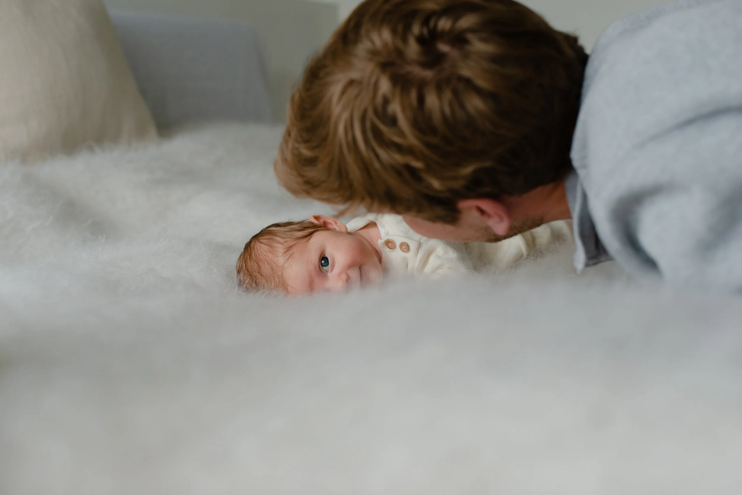 A man with brown hair lying on the floor face-to-face with a baby with reddish hair, on a light-colored fluffy surface.