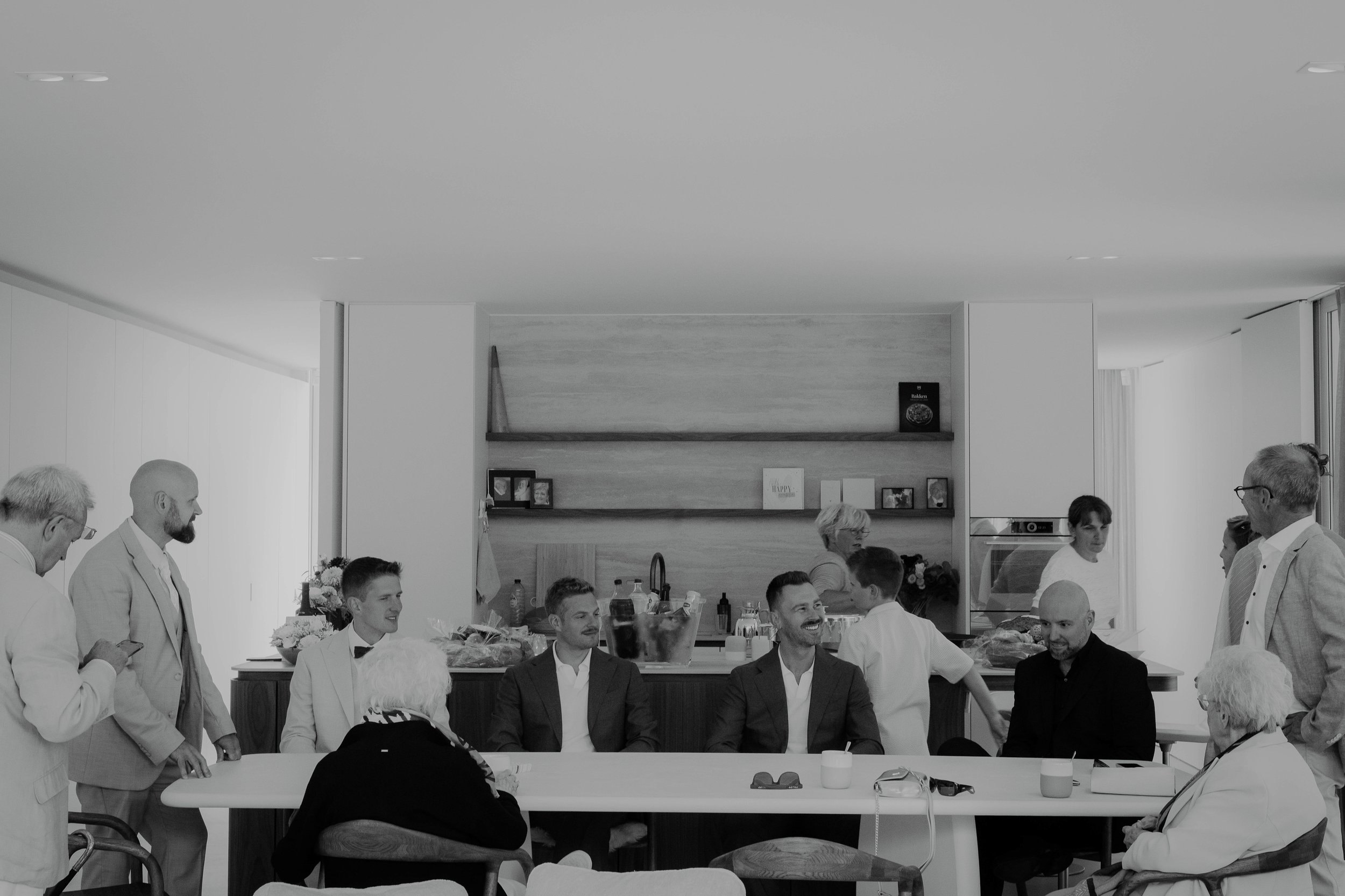 People gathered around a table in a bright, modern room, some engaged in conversation, with a kitchen area in the background.