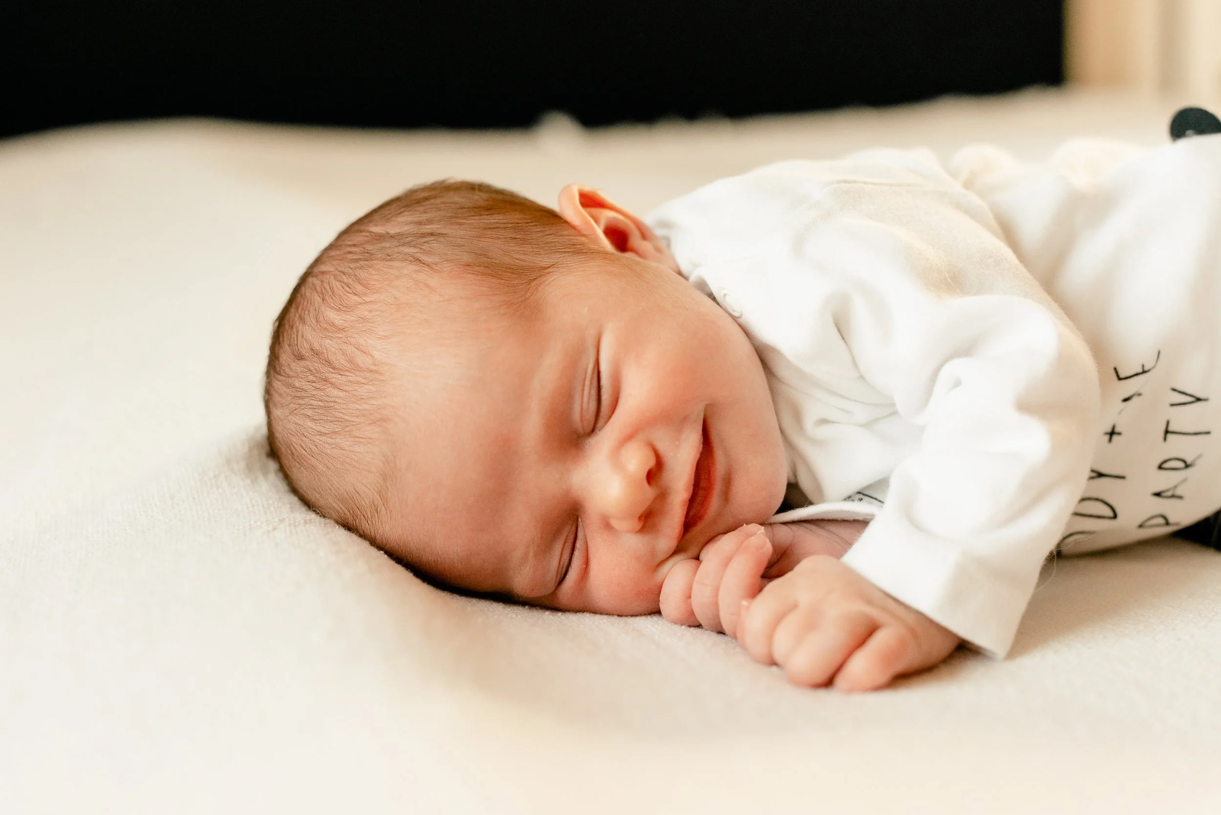 Close-up of a smiling newborn baby sleeping on a soft surface, wearing a white outfit.