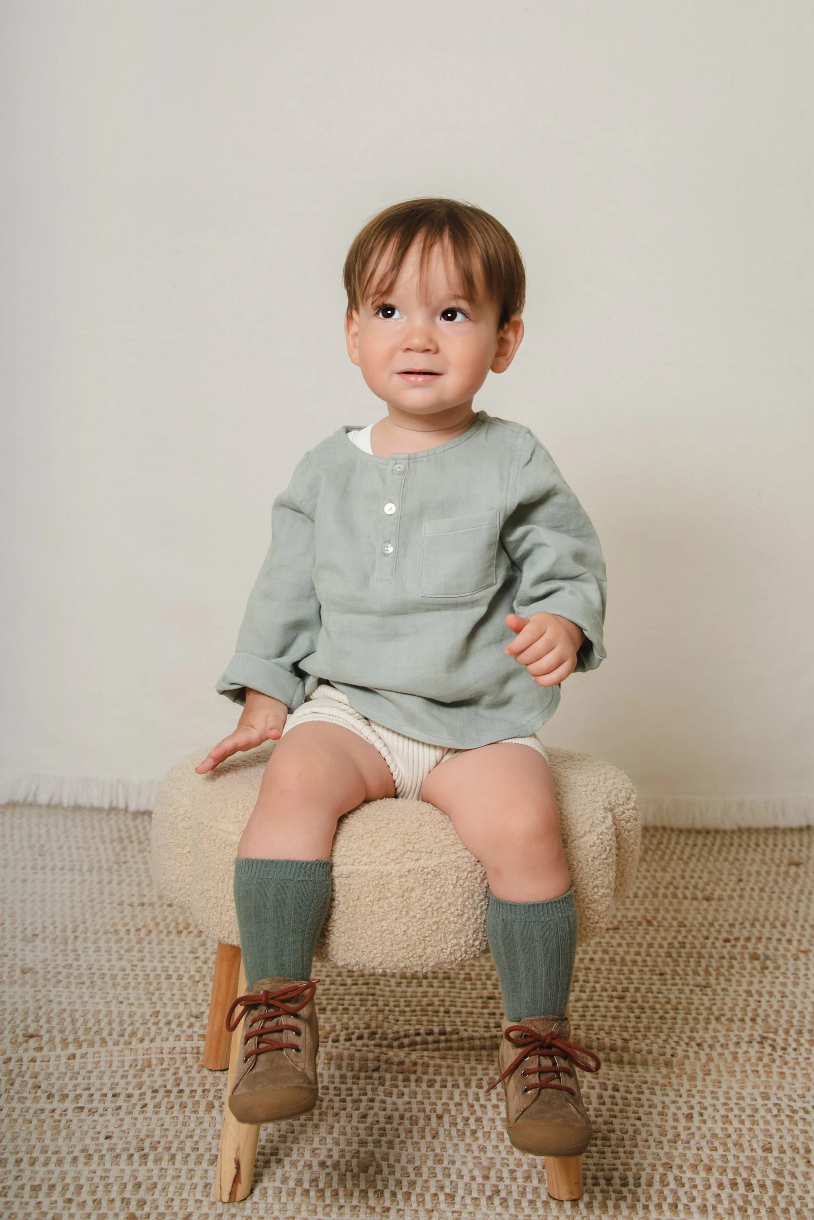 A young boy with brown hair sitting on a small beige upholstered chair, wearing a light green long-sleeved shirt, beige shorts, gray knee-high socks, and brown shoes.