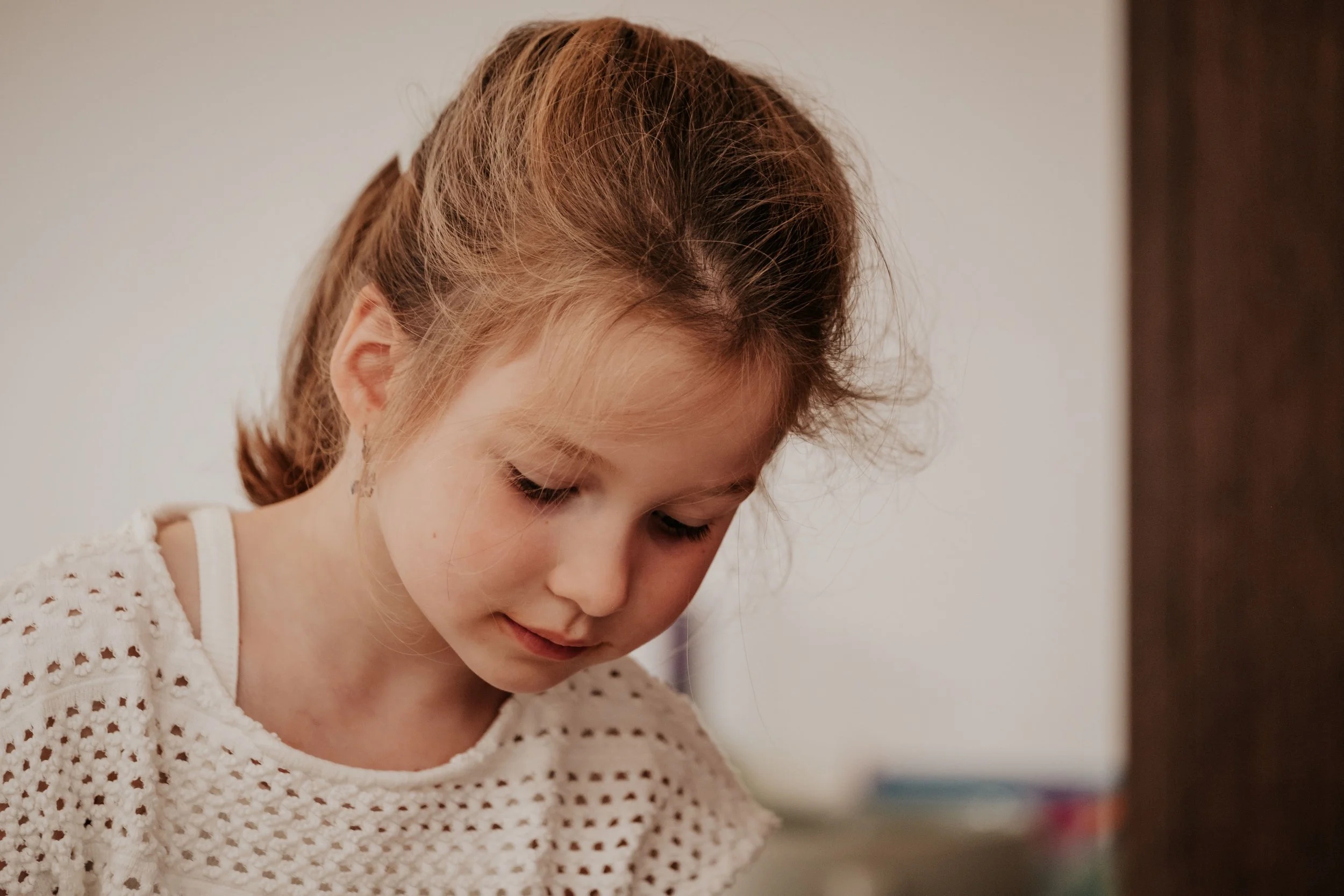 A young girl with light brown, curly hair wearing a white, perforated sweater, looking down.