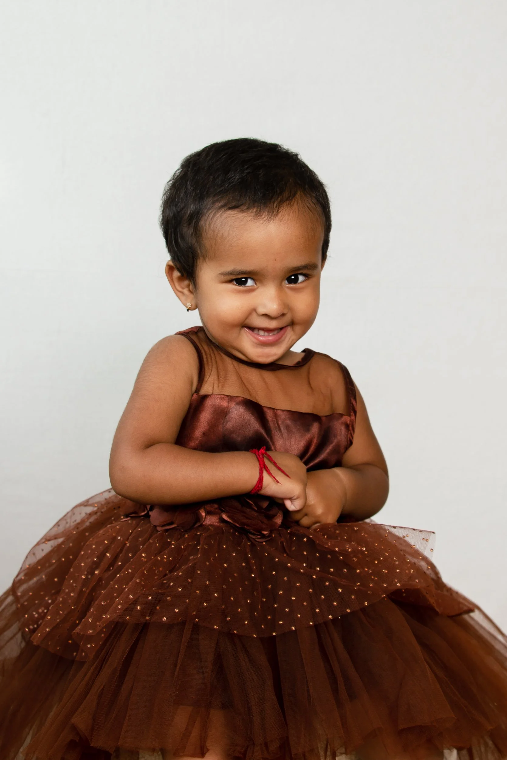 A young girl with short black hair and earrings is smiling and wearing a brown dress with a multi-layered, tulle skirt with gold dots, sitting against a plain white background.