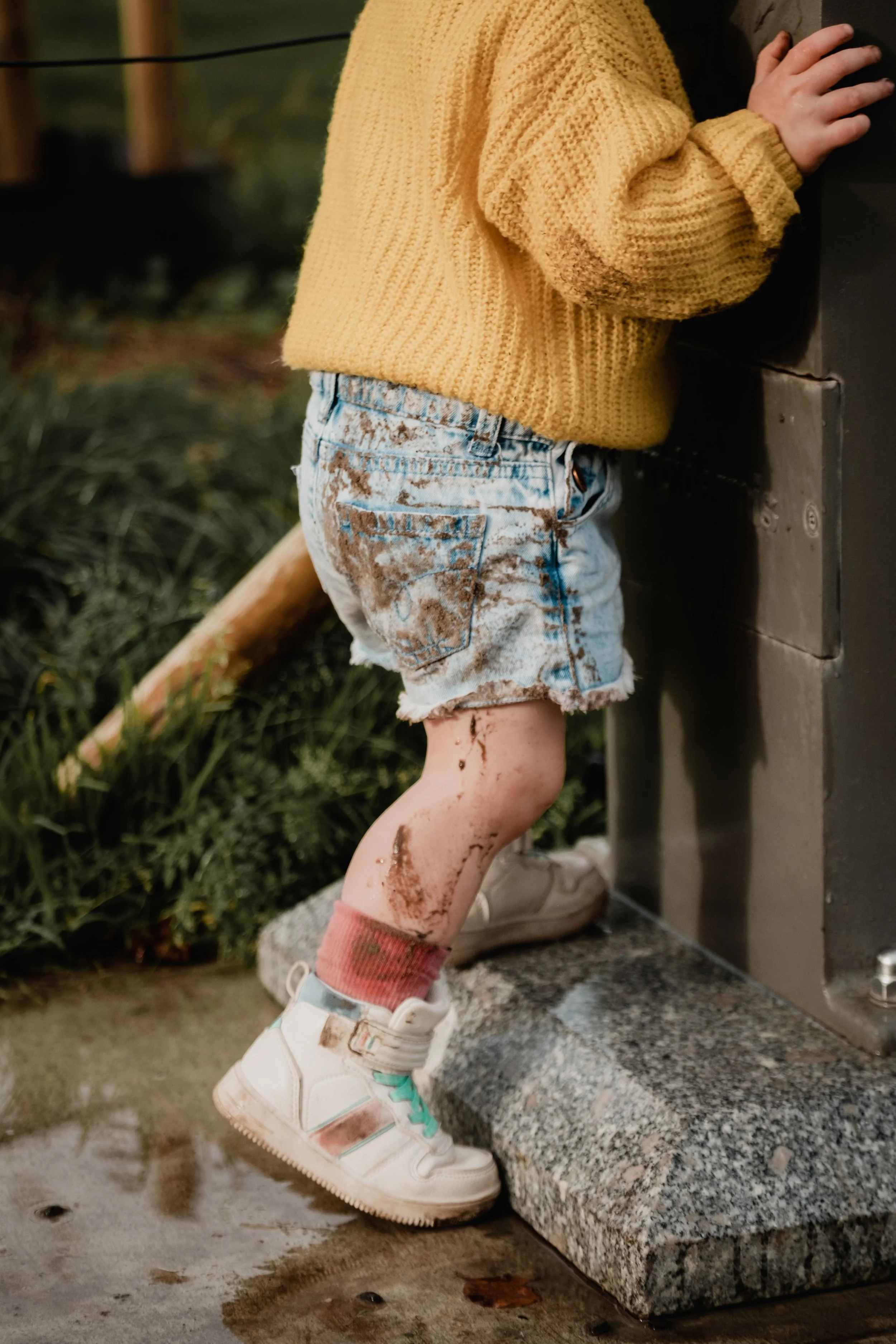Close-up of a child's muddy legs, wearing white sneakers, pink socks, and ripped denim shorts, standing on a concrete surface next to a bench with muddy hands resting on a dark surface, in an outdoor setting during evening or cloudy weather.