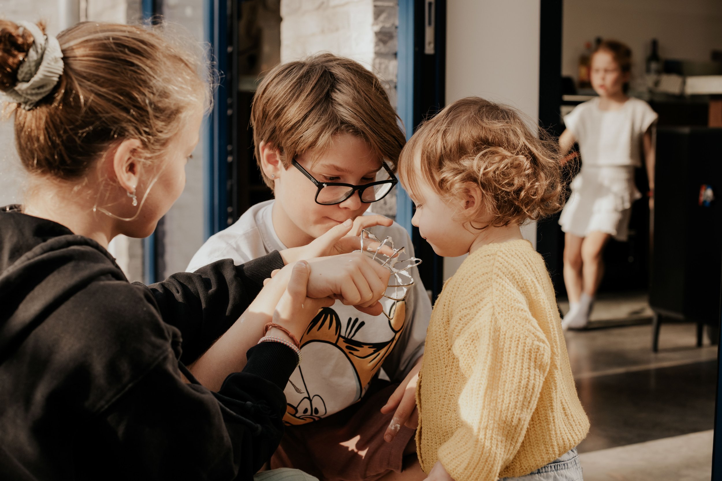Three children and one adult are examining an object together indoors, with a girl in a yellow sweater standing nearby and another girl in the background.