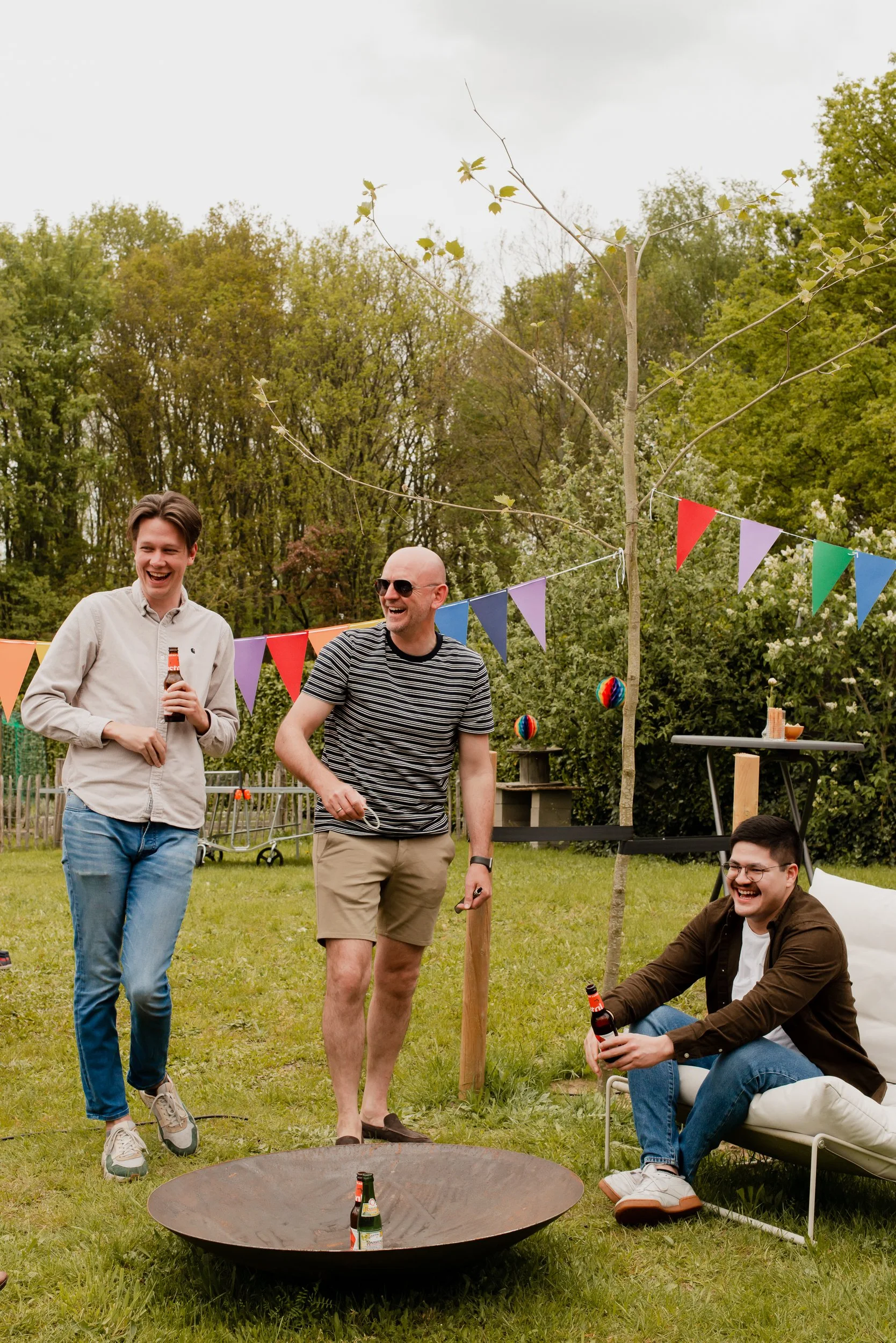 A group of four young men enjoying an outdoor gathering in a backyard with trees, bunting, and lawn games, some holding drinks and smiling.