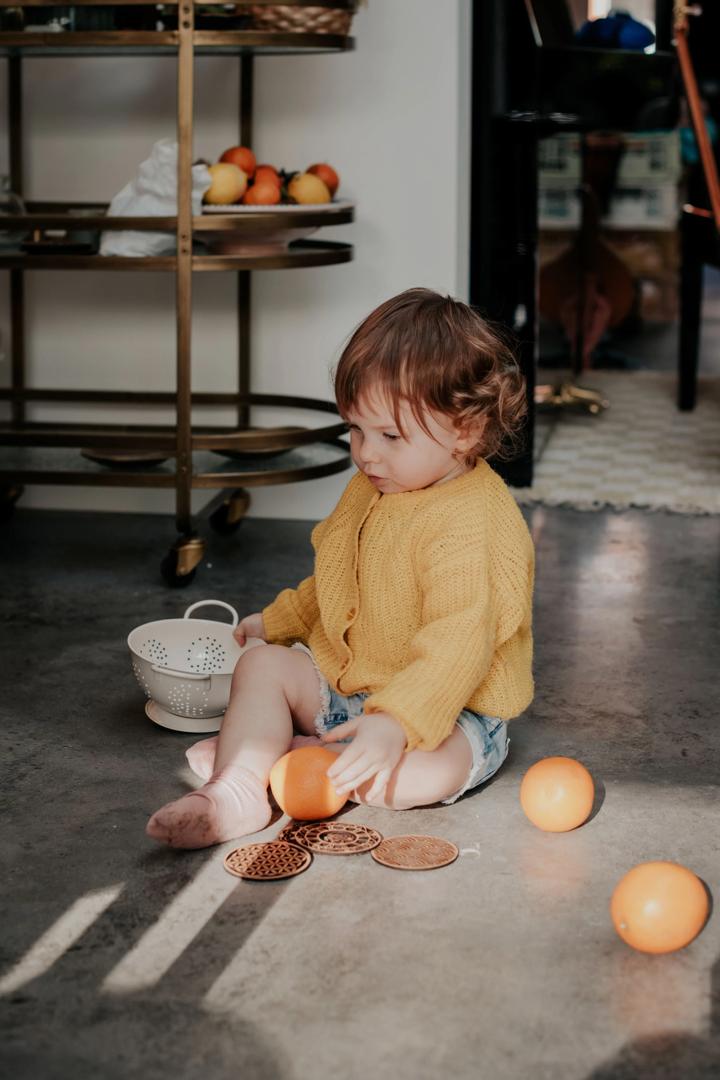 A young child with curly brown hair wearing a yellow sweater and denim shorts, sitting on a concrete floor, playing with orange objects and a white colander. Orange objects are scattered around, and some are in the child's hands. In the background, t