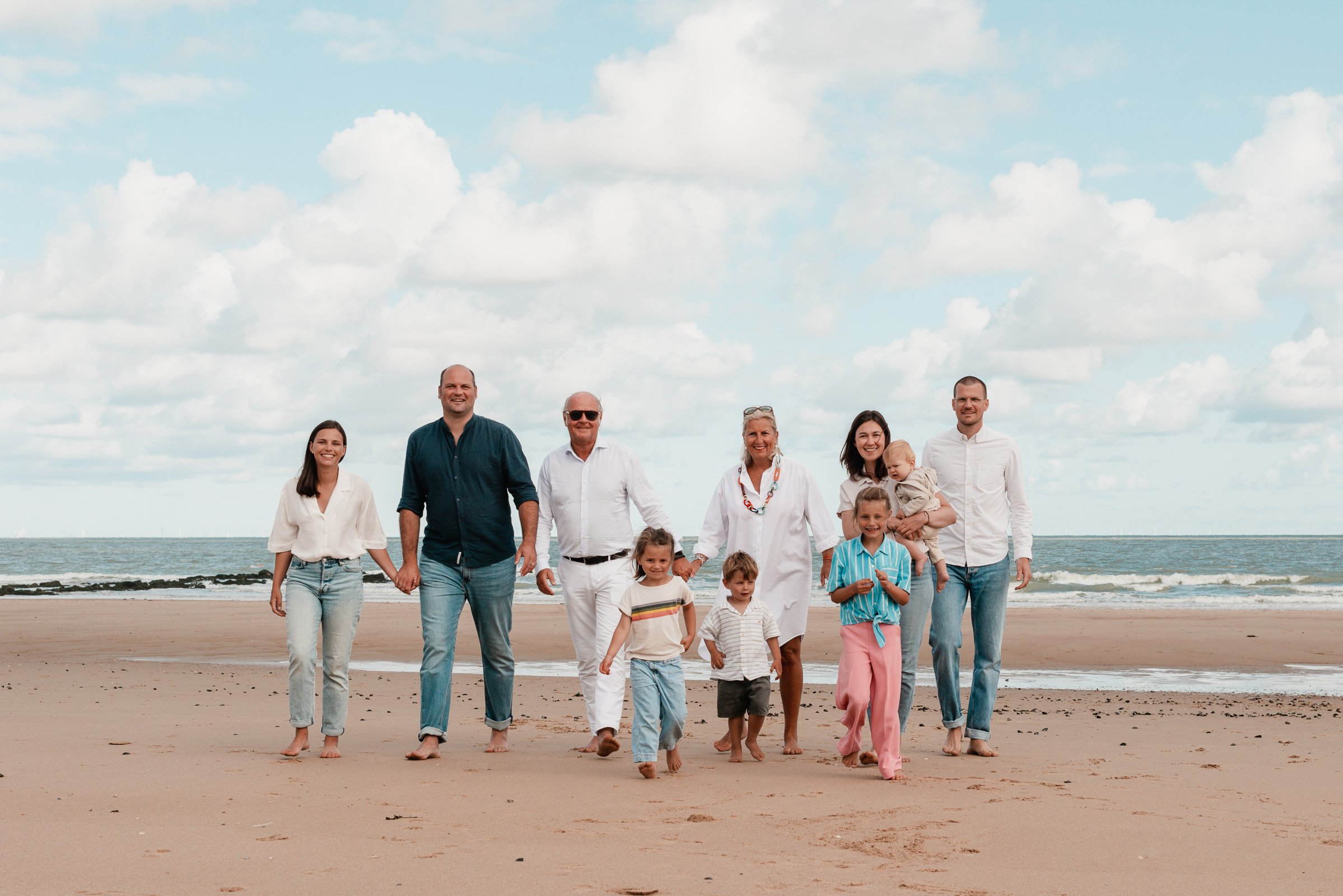 A large, multigenerational family walking along a sandy beach holding hands, with the ocean and a cloudy sky in the background.