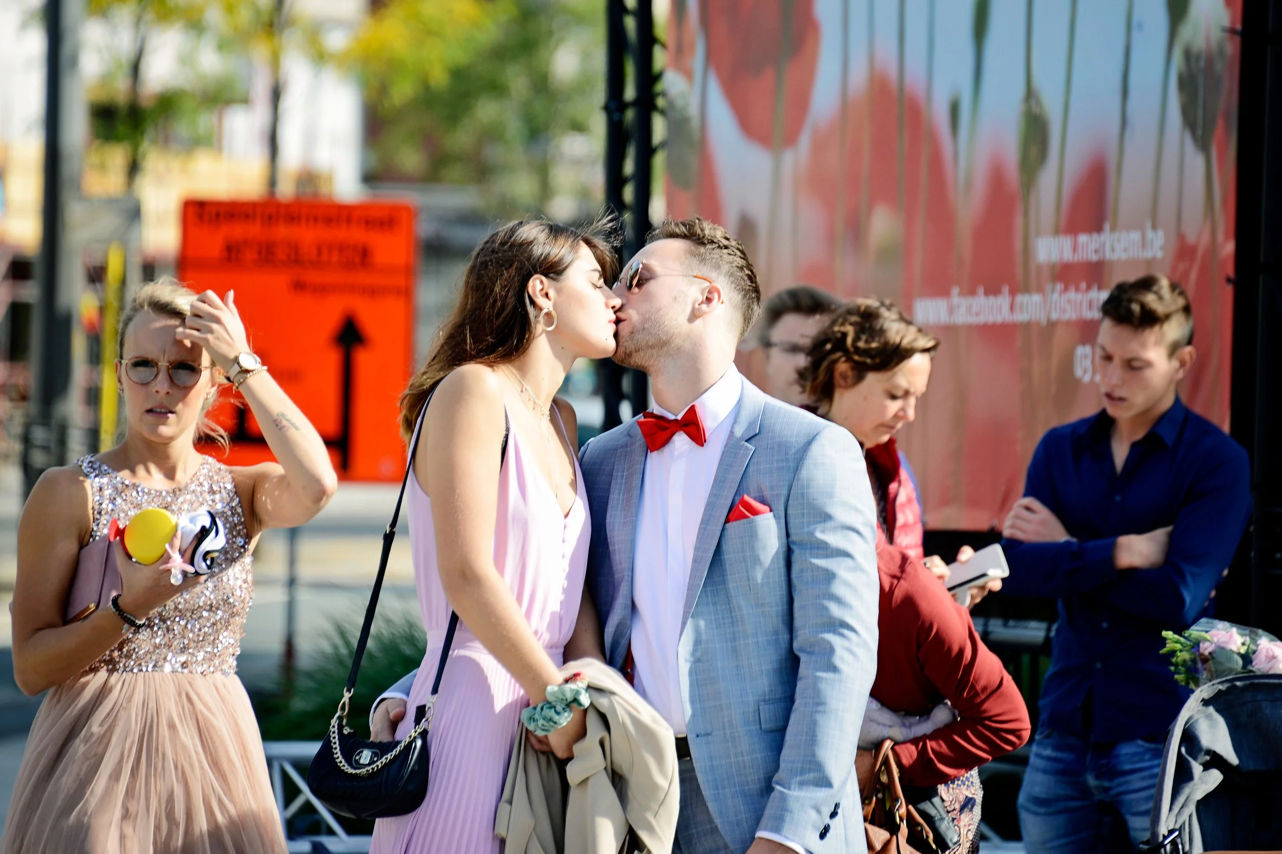 A couple is kissing in a crowded outdoor setting, with other people standing nearby, some looking at their phones. The man is wearing a light gray suit with a red bow tie and has sunglasses. The woman is wearing a pink dress with earrings. The backgr