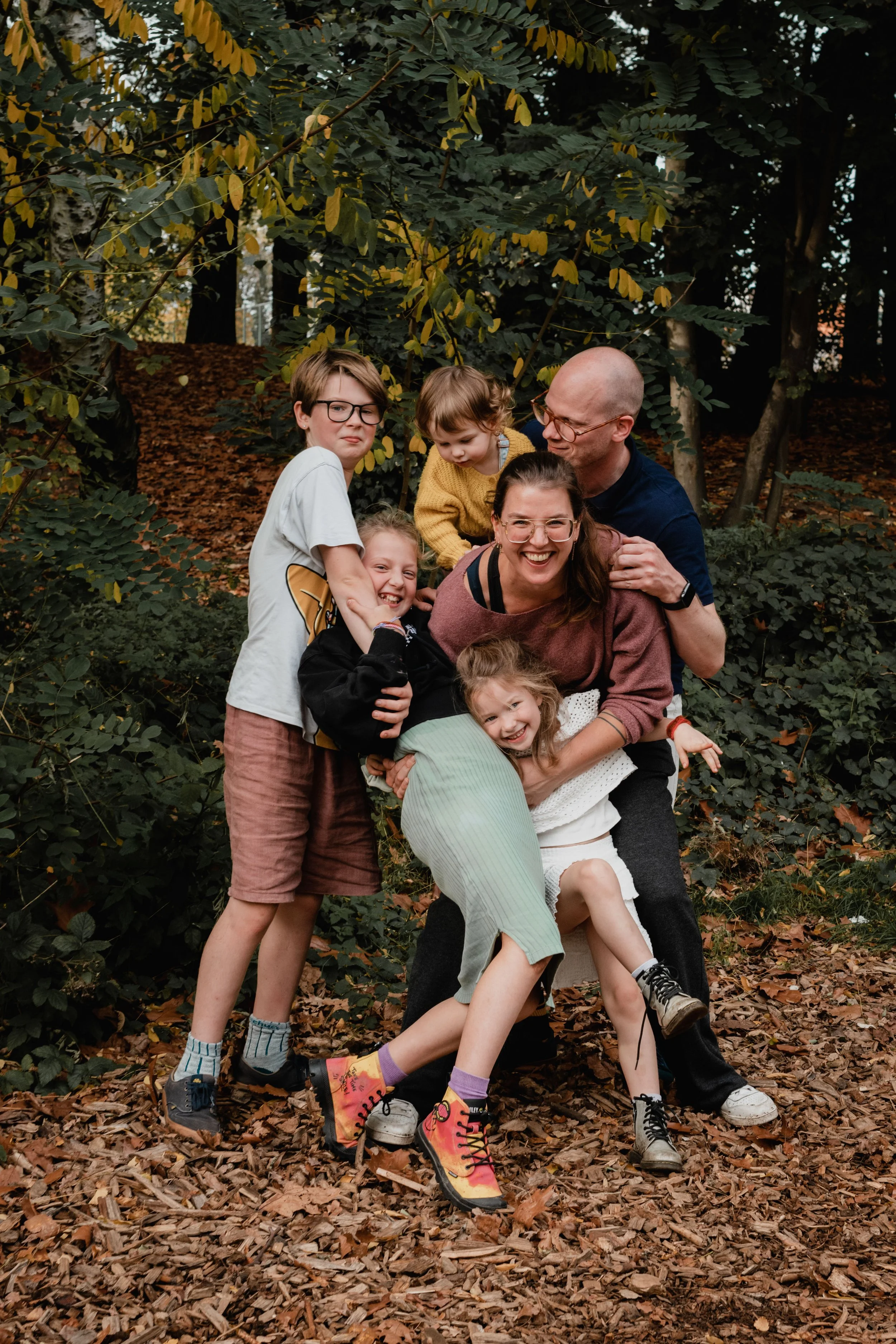 Family of six embracing outdoors in a wooded area with fallen leaves on the ground, smiling and playing.