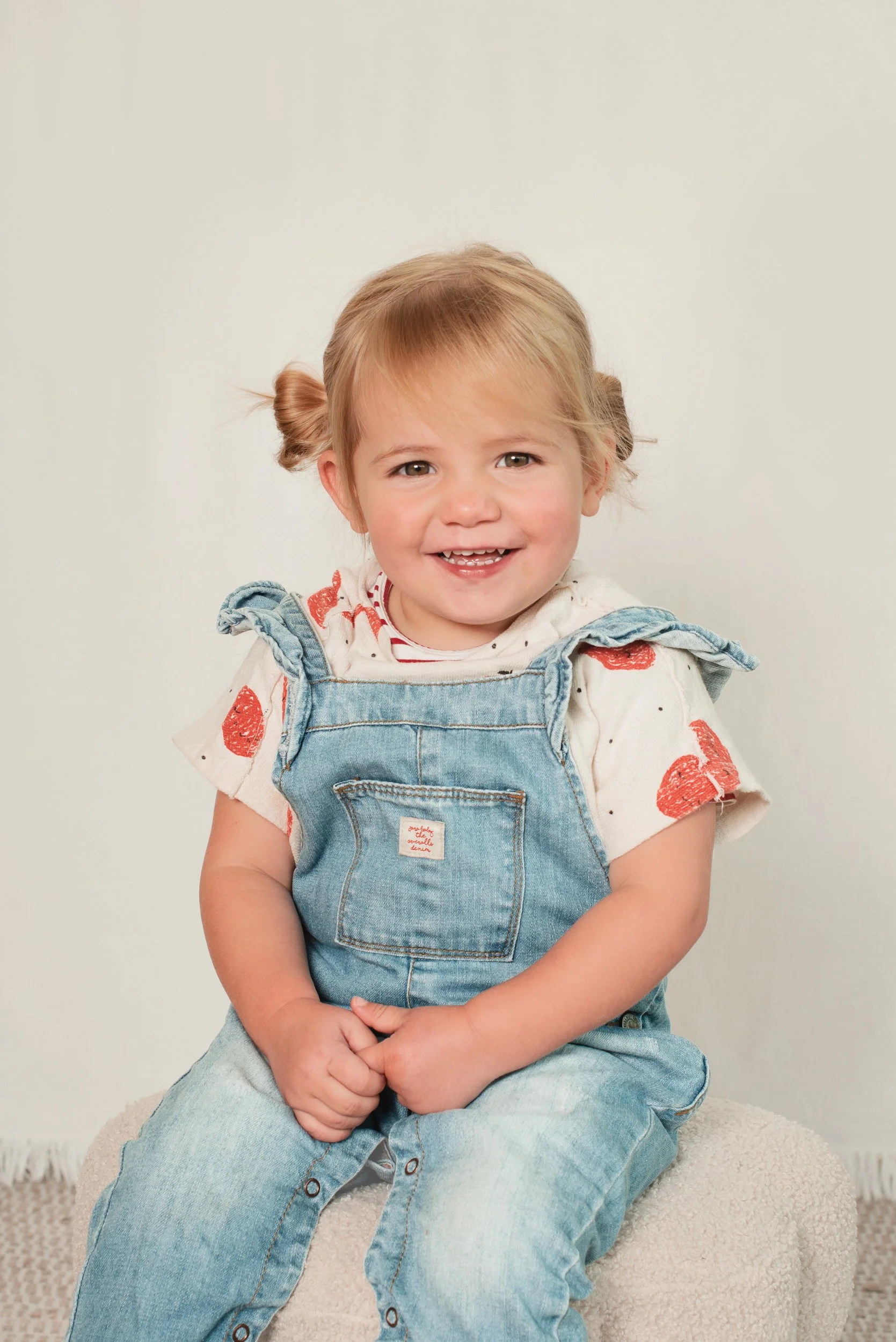 A young girl with blonde hair styled in two small buns, smiling with teeth visible, sitting on a cream-colored textured stool against a plain light background, wearing a white shirt with red design, denim overalls, and clasped hands resting on her la