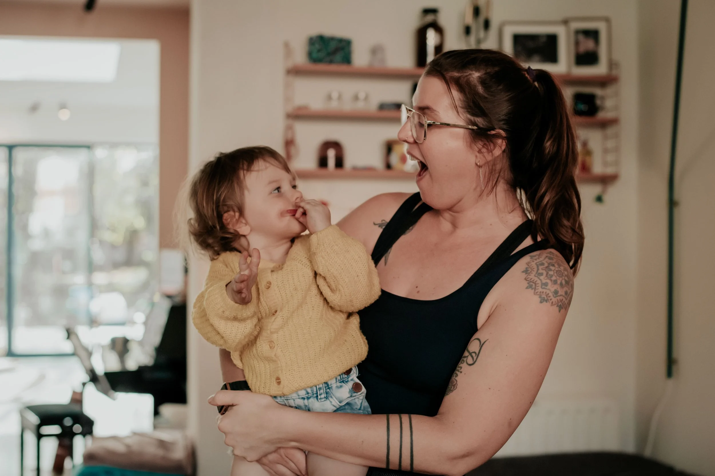 A woman with glasses smiling and holding a young girl in a yellow sweater in her arms inside a home, with shelves and framed pictures in the background.