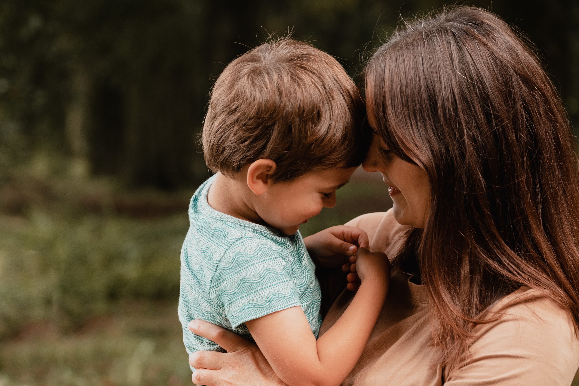 A woman with long brown hair holding a young boy close, both pressing their foreheads and noses together outdoors, with blurred trees in the background.