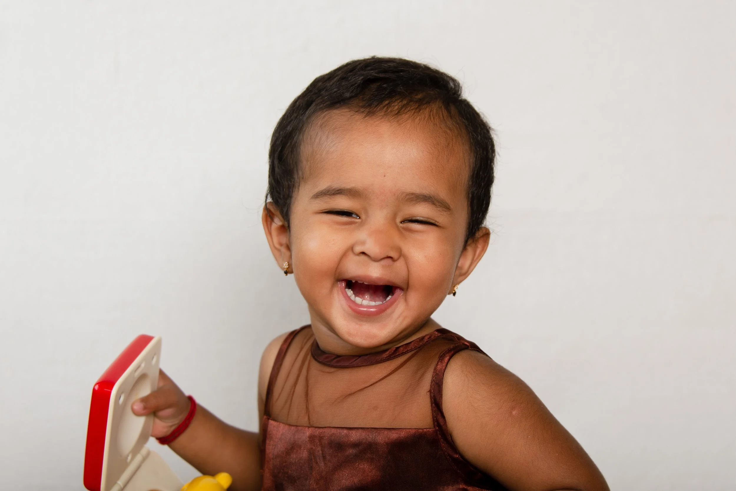 A young girl with short dark hair, wearing earrings and a brown dress with sheer fabric, laughing and holding a toy or object in her right hand, against a plain white background.