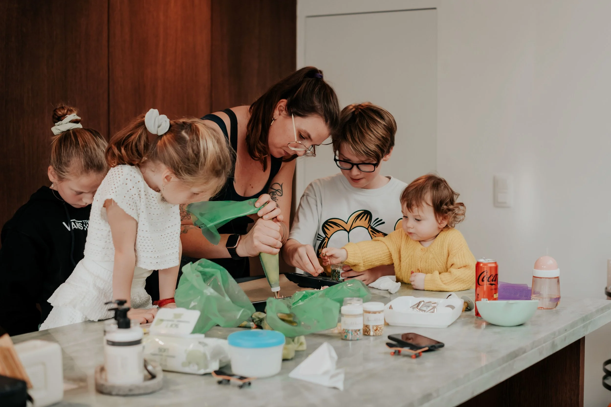 A woman with glasses and dark hair leaning over a table with four children, helping them with crafts or activities, with various supplies and drinks on the table.