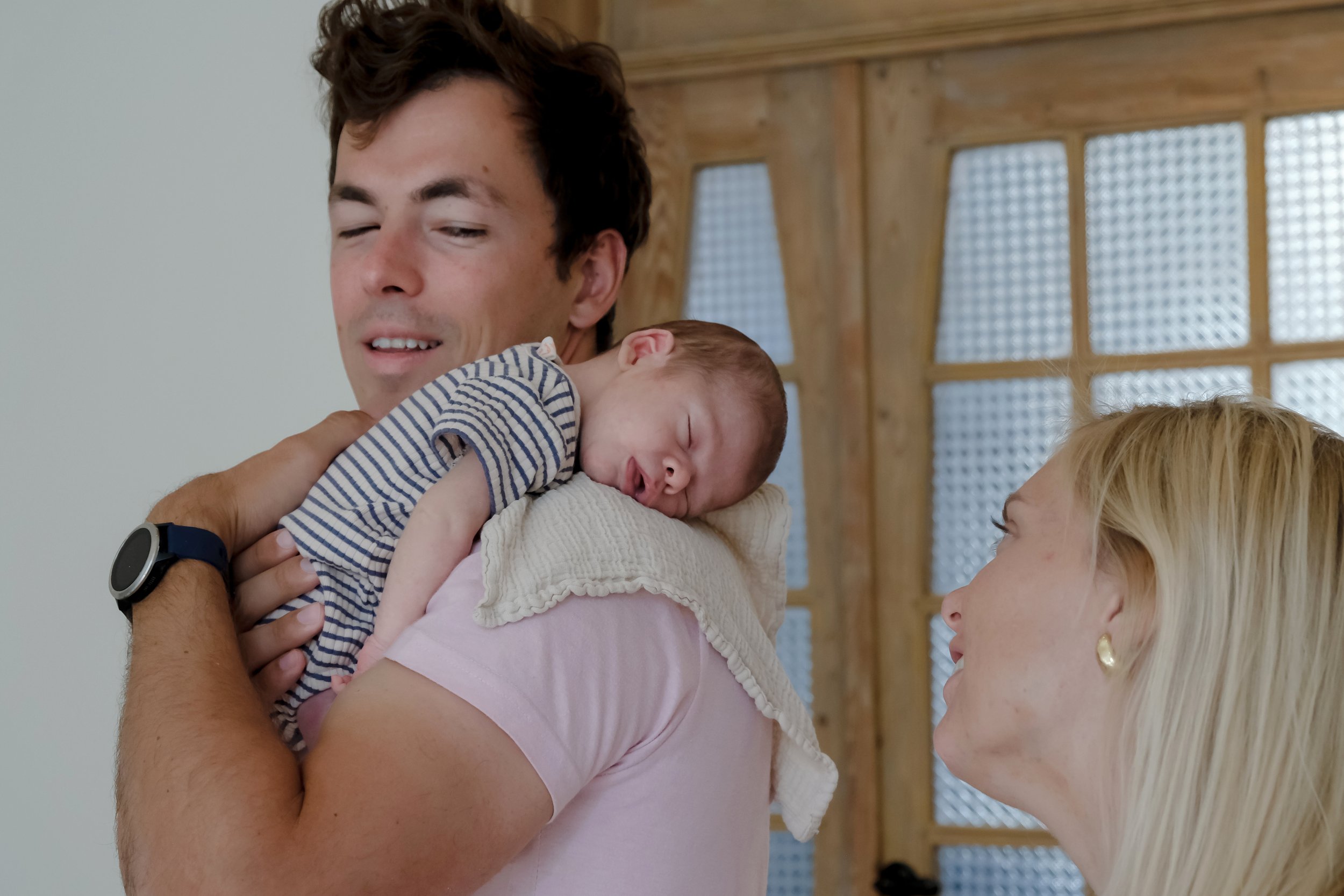 A man holding a sleeping baby in a room with wooden panel walls and glass block windows, while a woman with blonde hair looks at them.